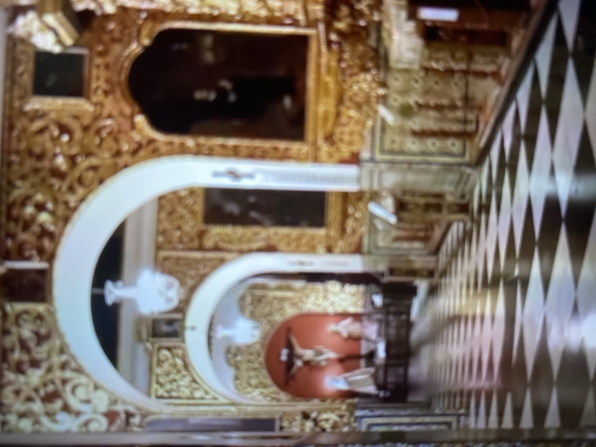 The image presents an upward view of a richly decorated interior, likely a religious building, specifically the Church of Tinaquillo in the city of Tinaquillo, Venezuela.
The scene is structured around a series of arches, which are painted white and lead the eye into the depth of the space. The surrounding architecture is heavily adorned with gold-colored ornamentation. One distinct feature is a black and white checkered floor, running diagonally across the right side of the image, adding a contrasting geometric element.
At the end of the archway, a crucifix is visible, further indicating the religious context. The lighting is unclear, but the focus seems to be on the crucifix, suggesting it's a focal point within the interior.