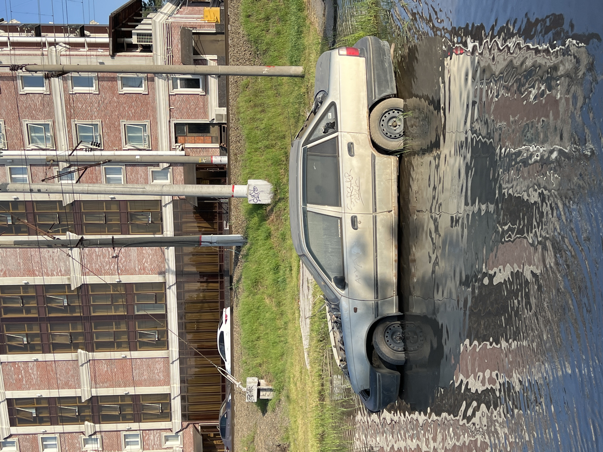 The scene depicts a beige-colored, abandoned car partially submerged in water, possibly floodwater, next to a grassy area adjacent to a building in Krasnodar, Russia. The building appears to be made of brick with multiple windows. There is evidence of graffiti or markings on the side of the car. Electrical poles and wires are visible in the background. The water's surface is rippled, reflecting the car and the surrounding environment. Two other vehicles are visible on the left of the frame.