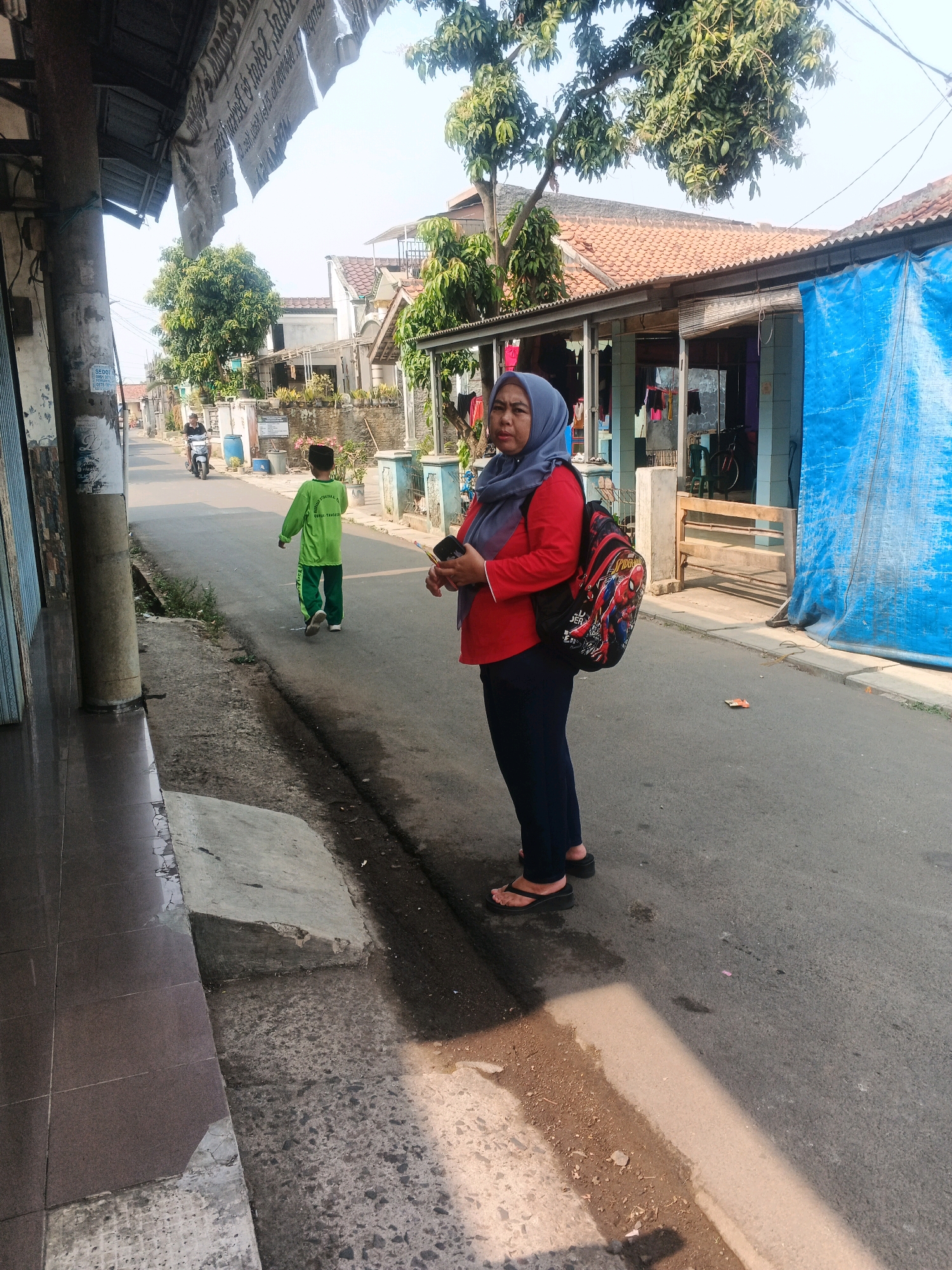 An Indonesian woman wearing a red shirt, blue headscarf, and black pants stands on a paved road in Kadu, Indonesia. She carries a backpack with a Spider-Man design. A young boy in a bright green uniform walks away from her down the street. In the background, a motorbike is visible in the distance. The scene appears to be a residential area, with houses and trees lining the road. The sky is bright and clear, suggesting a sunny day.