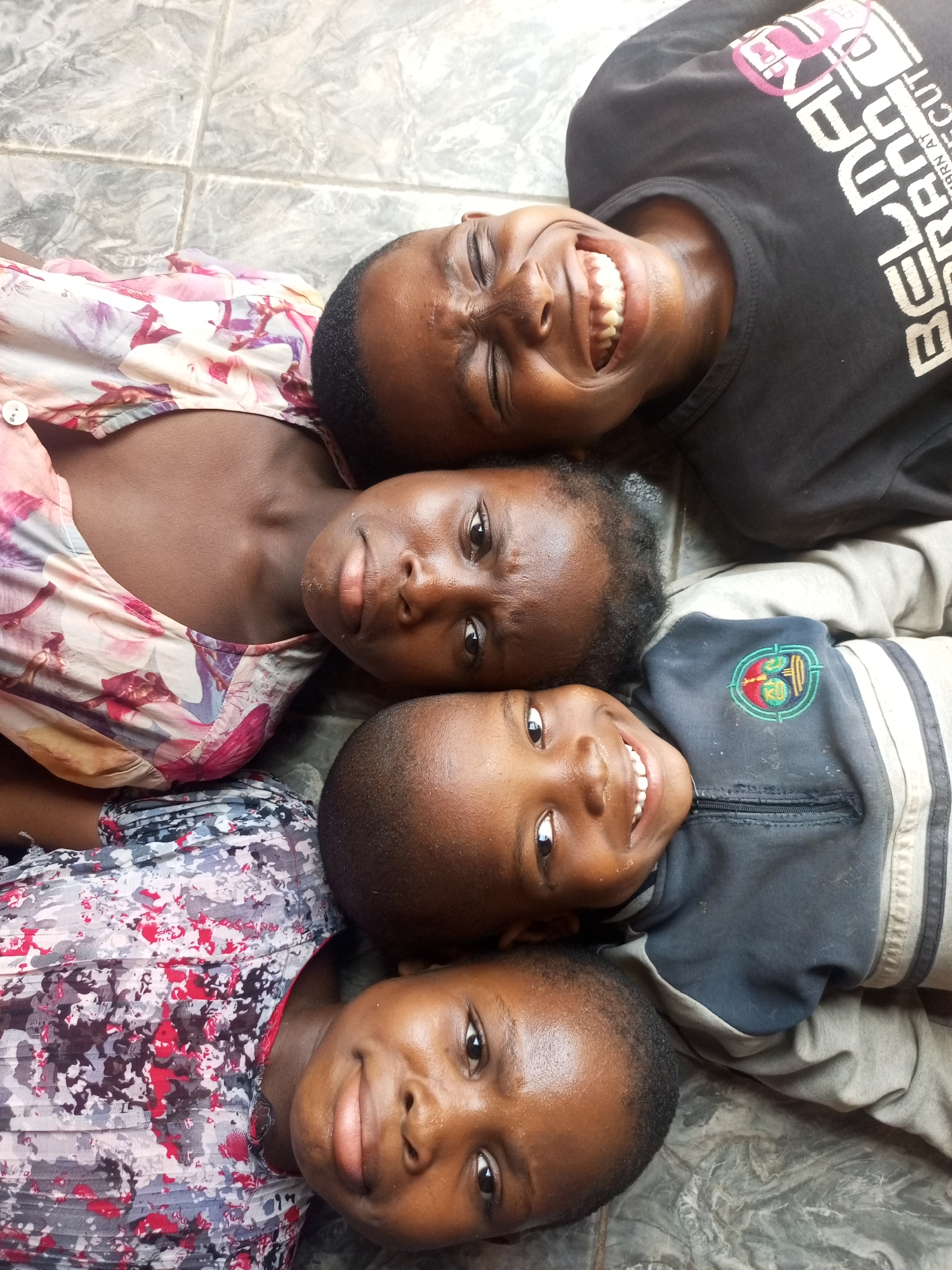 Four Black children lie on a tiled floor, heads oriented towards the center, in Tempure, Nigeria. From top to bottom: a teenager wearing a black t-shirt smiles widely. Next, an adolescent wearing a floral print top looks at the camera with a slight frown. Below, a younger child wearing a blue and grey zip-up smiles. At the bottom, a child wearing a dark patterned top looks towards the camera with a soft smile. The background appears to be an interior space with light-colored tiling.