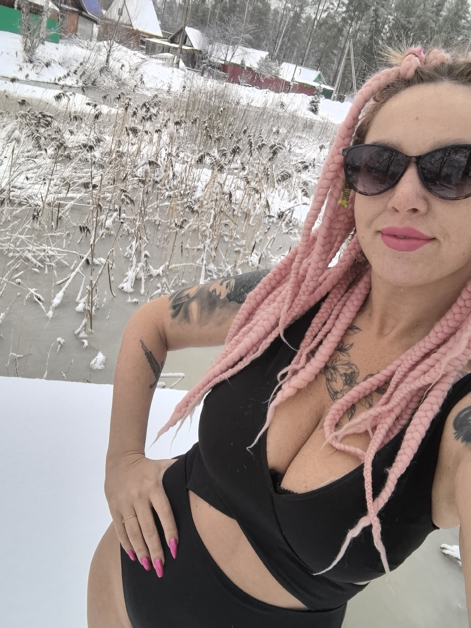 A selfie-style photograph shows a woman with pink braided hair and sunglasses standing in a snowy landscape. She is wearing a black swimsuit-like garment and pink nail polish. The woman has tattoos on her arms and chest. The background features a body of water or frozen river with snowy banks and vegetation. In the distance, there are residential buildings. Based on the prompt, the location is identified as Noviy, Russia.