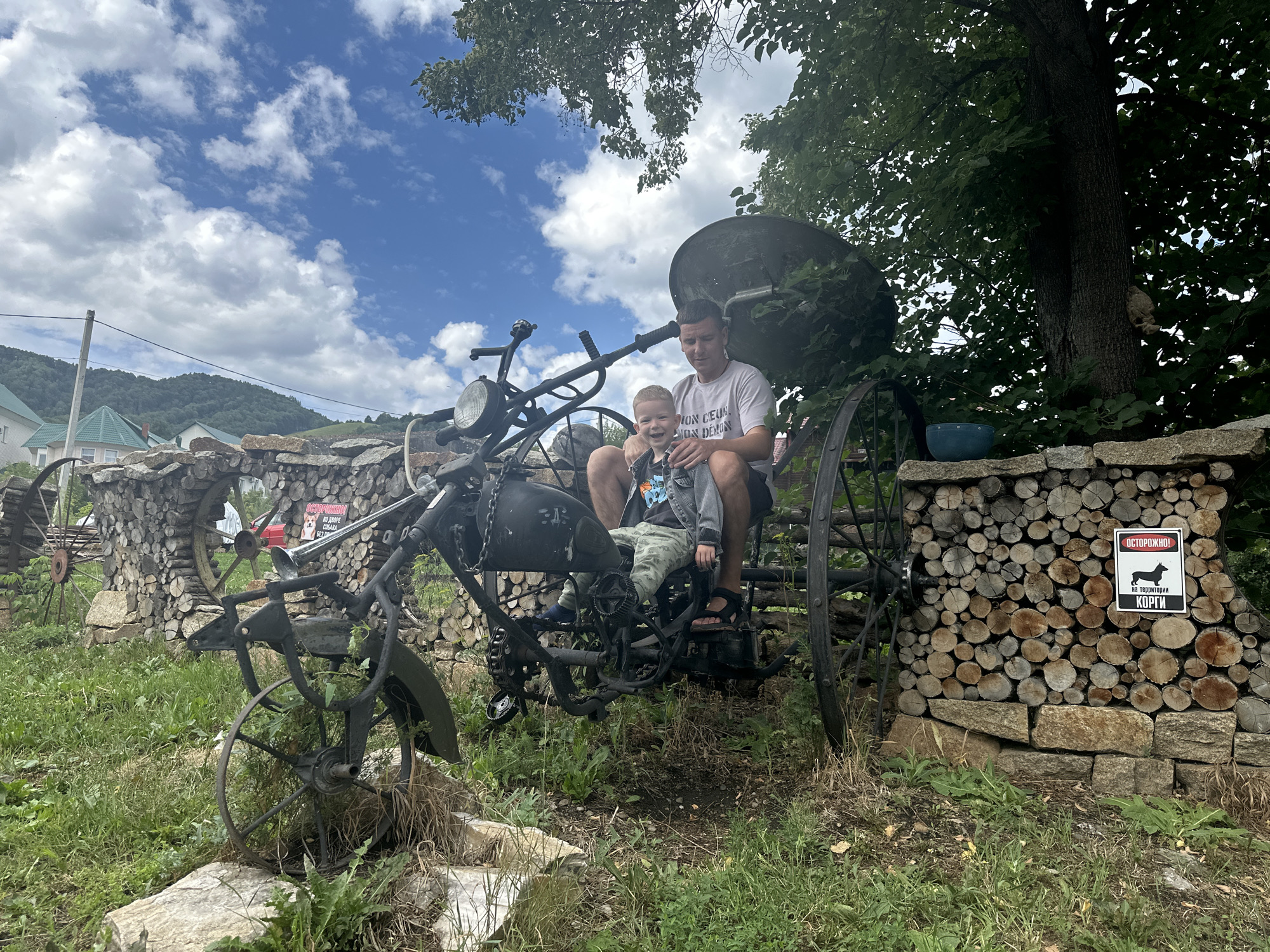 An outdoor shot shows a man and a young boy sitting on a custom-made, rustic motorcycle-like sculpture. The man is seated behind the boy, with his arm around him. Both are smiling. They are dressed in casual clothing. The sculpture is dark, made of metal and what appears to be repurposed parts, with large spoked wheels. It is positioned in front of a wall constructed from stacked logs and stones, with a sign on it that reads "ОСТОРОЖНО!" (Caution!) and depicts a corgi. The background features a lush green hillside with houses and trees, under a blue sky with white clouds. The overall setting suggests a rural or semi-rural environment, possibly a yard or a park in Даниловка (Алтайский), Russia. The foreground is covered in green grass and some stones.
