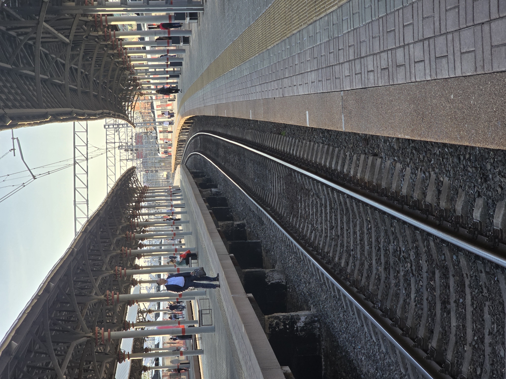The image shows a train platform in Moscow, Russia, likely at one of the larger stations. The platform is covered by a metal structure supported by vertical pillars. Passengers can be seen on the platform. The train tracks run alongside the platform, with a noticeable curve in the tracks. The platform's edge is marked with a textured strip for safety. The weather appears to be clear and sunny. The image is taken from a slightly elevated perspective.