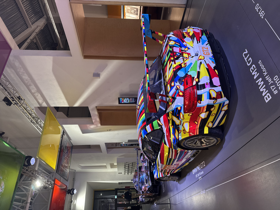The image features a vividly painted BMW M3 GT2, adorned with a vibrant and abstract design. Text beside it reads "BMW M3 GT2 #17 Jeff Koons 2010". The car is positioned in what appears to be a museum or exhibition space, with the colorful artwork on the car contrasting against the neutral background. The composition emphasizes the car's unique design and artistic significance. Additional cars appear in the background, lined up in the exhibition.