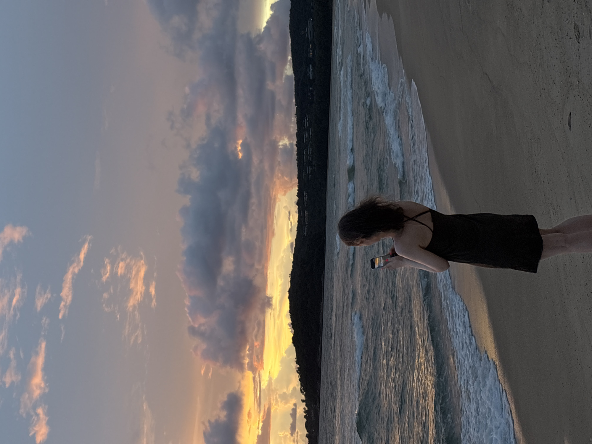 A person wearing a black dress stands on a sandy beach in Choeng Thale, Thailand, facing the ocean and holding a smartphone to take a picture. The ocean has moderate waves, and the sky is filled with dramatic clouds illuminated by a sunset, with hues of orange, yellow, and purple. A dark, tree-covered headland is visible in the distance across the water. The lighting suggests it is late afternoon or early evening.
