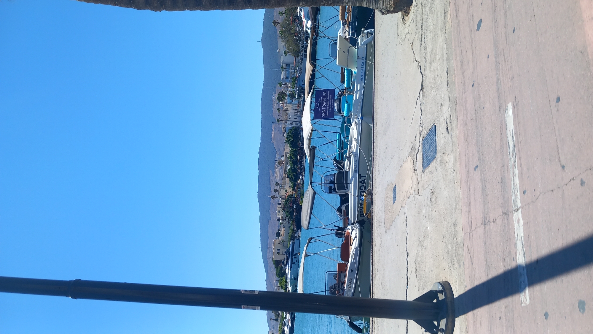 The image shows a waterfront scene in Kos, Greece, with several small boats docked along a concrete pier. In the foreground, a dark pole casts a shadow across the pier and a pinkish-red paved area. Behind the pole, the blue water of the harbor is visible, with several boats moored. The boats are of various sizes and appear to be for rent, with awnings providing shade. Some of the boats have text on them, including "RENT BOAT" and what appears to be Greek lettering. In the background, a town or city is visible with buildings and hills beyond. The sky is clear and blue. The overall scene depicts a sunny day at a harbor, likely a tourist area, given the rental boats and the visible town.