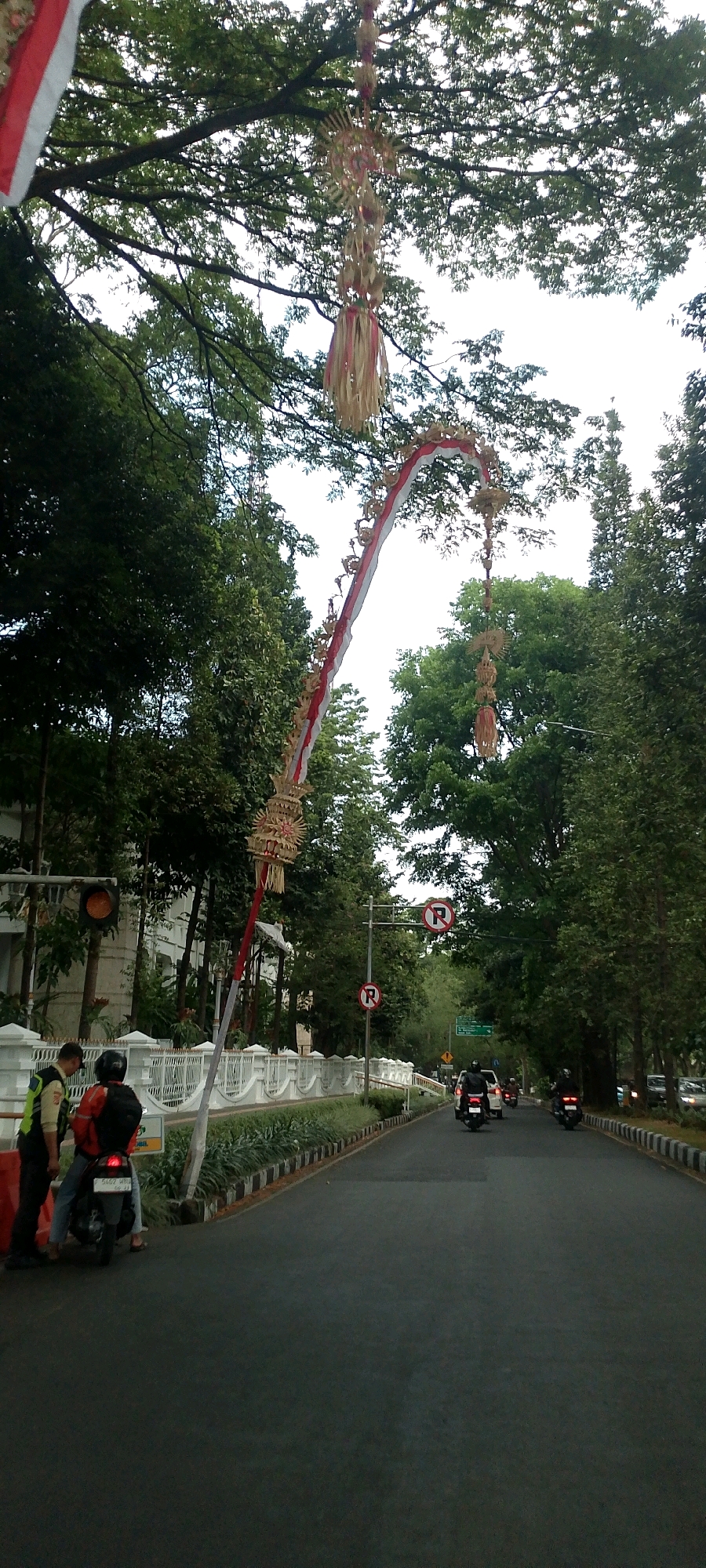 The image depicts a street scene in Bandung City, Indonesia, likely during daytime given the lighting. A prominent feature is a tall, arching decorative banner, characteristic of Balinese cultural decorations known as a "Penjor," which extends across the road from the left. This Penjor is adorned with woven palm leaves and ribbons, and its base is near a white, ornamental fence. Two individuals are standing next to a parked scooter on the left side of the road. One person, wearing a reflective vest, appears to be interacting with the scooter rider who is wearing a red jacket and a helmet.
The road is asphalt and appears to be a main thoroughfare with vehicles, including several motorcycles and a car, visible in the distance. Traffic signals are present, with at least one signal showing an amber light. Several "No Parking" signs are also visible on poles along the road. The environment is lined with lush green trees, suggesting a park-like or tree-lined avenue. Buildings are visible on the left side behind the white fence, appearing to be of modern architectural style. The sky is overcast, creating diffused lighting conditions. The presence of the Penjor suggests a possible cultural event or festival taking place in the area.