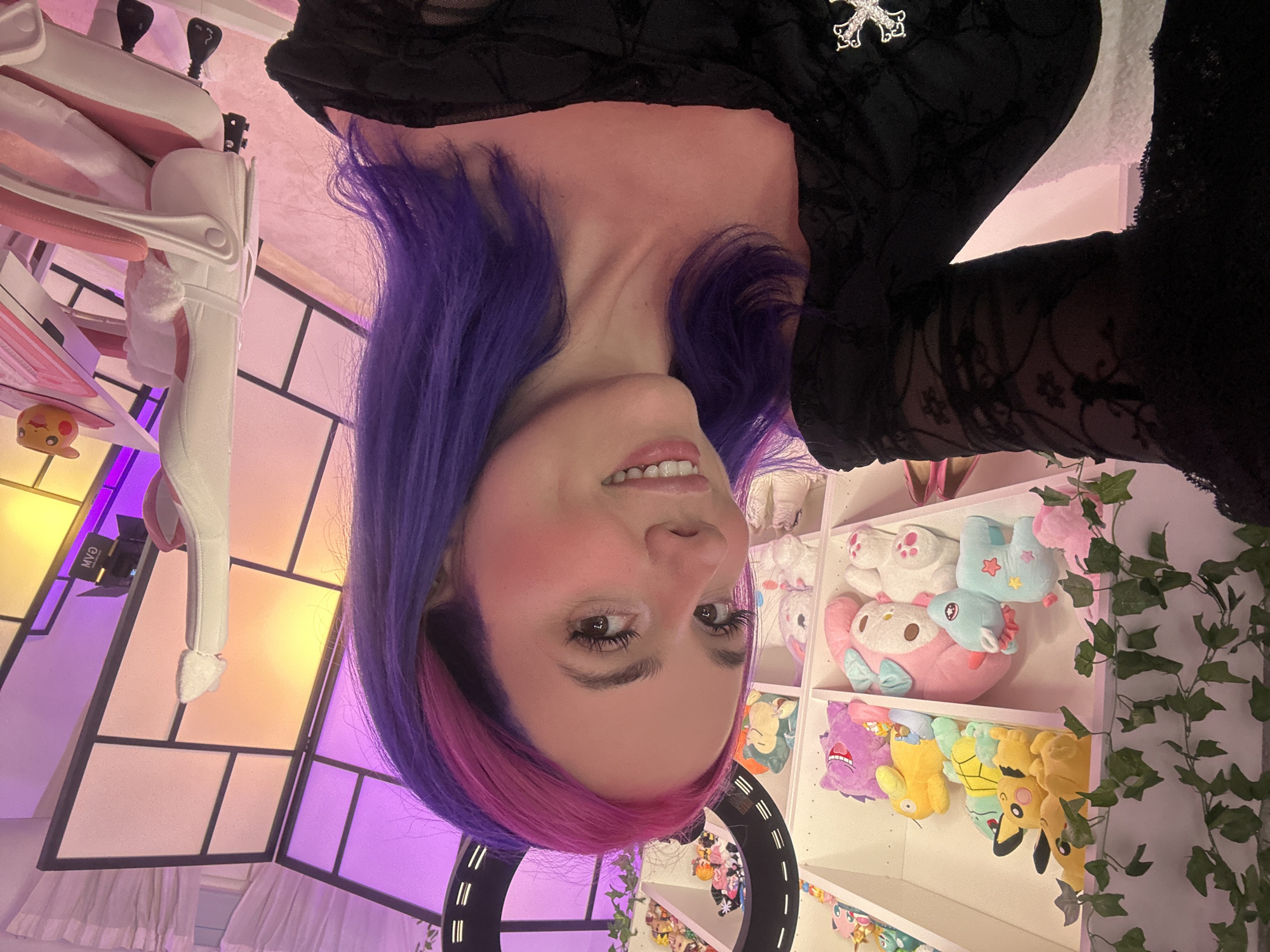 The image shows a young woman with vibrant purple and pink hair, wearing a black off-the-shoulder top with lace detailing and a silver cross brooch. She has makeup on and is smiling. She is positioned in what appears to be a room decorated with anime-themed plush toys on shelves, including Pokemon characters like Pikachu and Squirtle, and Sanrio characters like My Melody. A ring light is visible behind her. The room is lit with purple and pink ambient lighting. There is also a white and pink gaming chair with fluffy armrests, and a desk with a small Pikachu figurine. The background features a large light panel with a grid design. The scene is consistent with a streamer's setup or a themed room. There is no indication of the location being in the city of Oviedo, Spain.