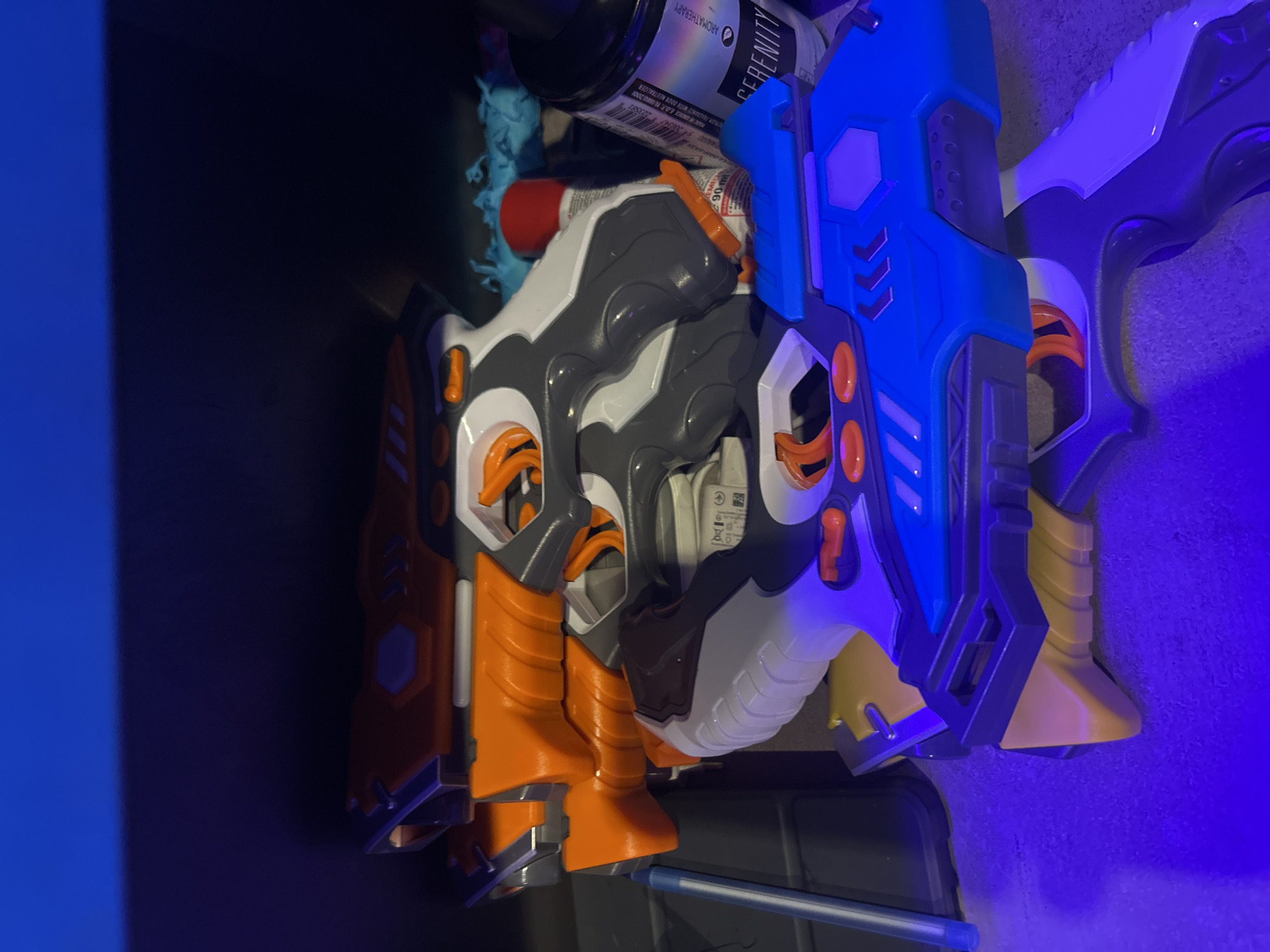 The image shows a collection of toy guns stacked on a shelf. The guns are various colors, including orange, gray, white, and blue. Some of the guns have orange triggers and detailing. In the background, there are some blue plastic toys, a red object, and a black bottle with text on it. The shelf appears to be dark, possibly made of wood, and is lit with blue and purple lights. The overall scene is a close-up shot of the toy guns. There is no indication of the city of Burgas, Bulgaria.
