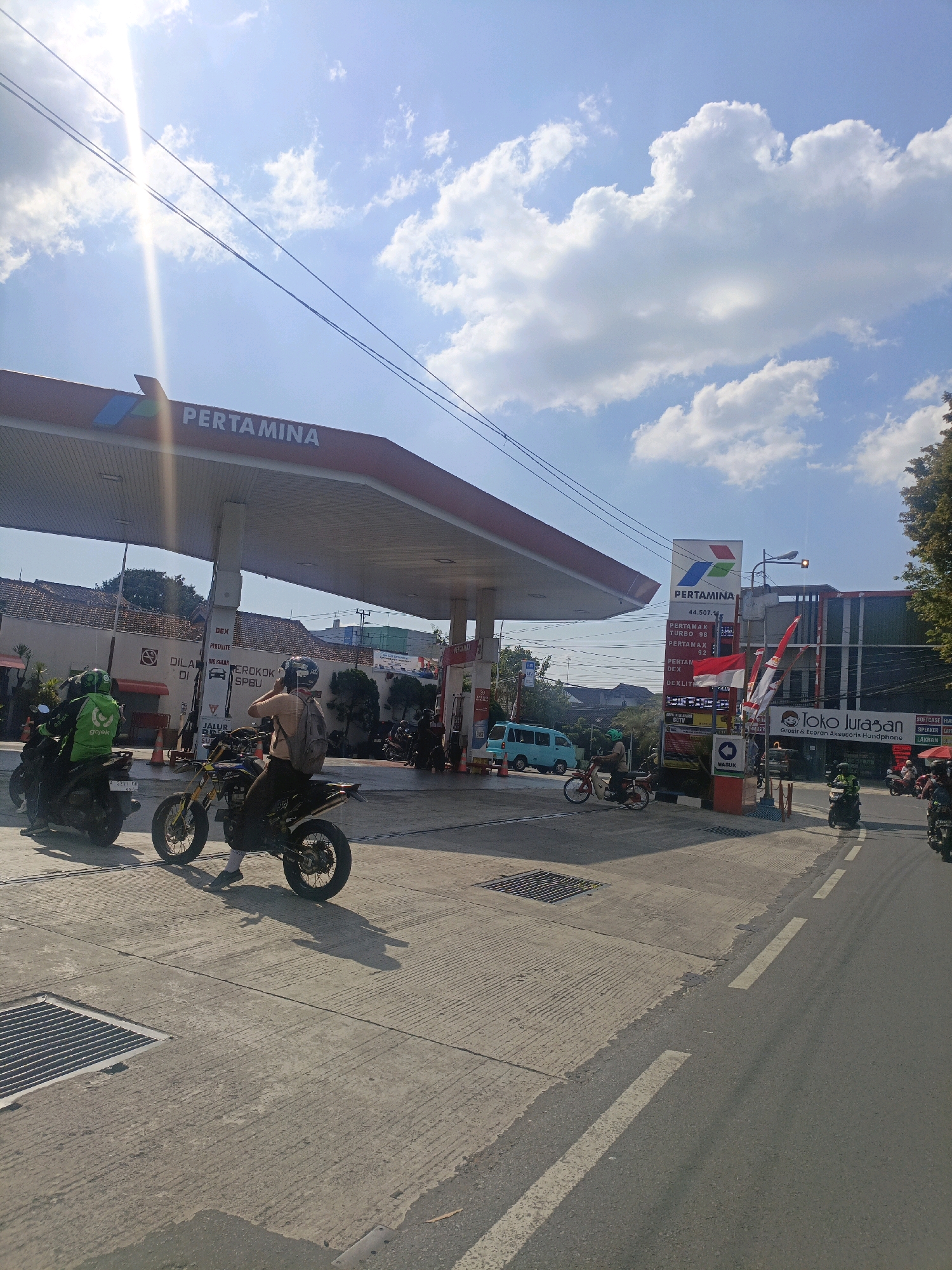 The image depicts a sunny day at a Pertamina gas station in Salatiga, Indonesia. A red and white Pertamina sign is prominently displayed, along with fuel pumps. Several motorcycles are visible, including one with a rider wearing a green Gojek uniform and another with a rider in a yellow shirt and backpack, both parked or in motion on the pavement. A light blue van is parked on the left side of the road. Buildings with commercial signage, including "Toko Juragan," are located behind the gas station. The sky is blue with scattered white clouds, and power lines are visible overhead. The ground is paved with concrete and asphalt, and a metal grate is present on the left side of the pavement.
