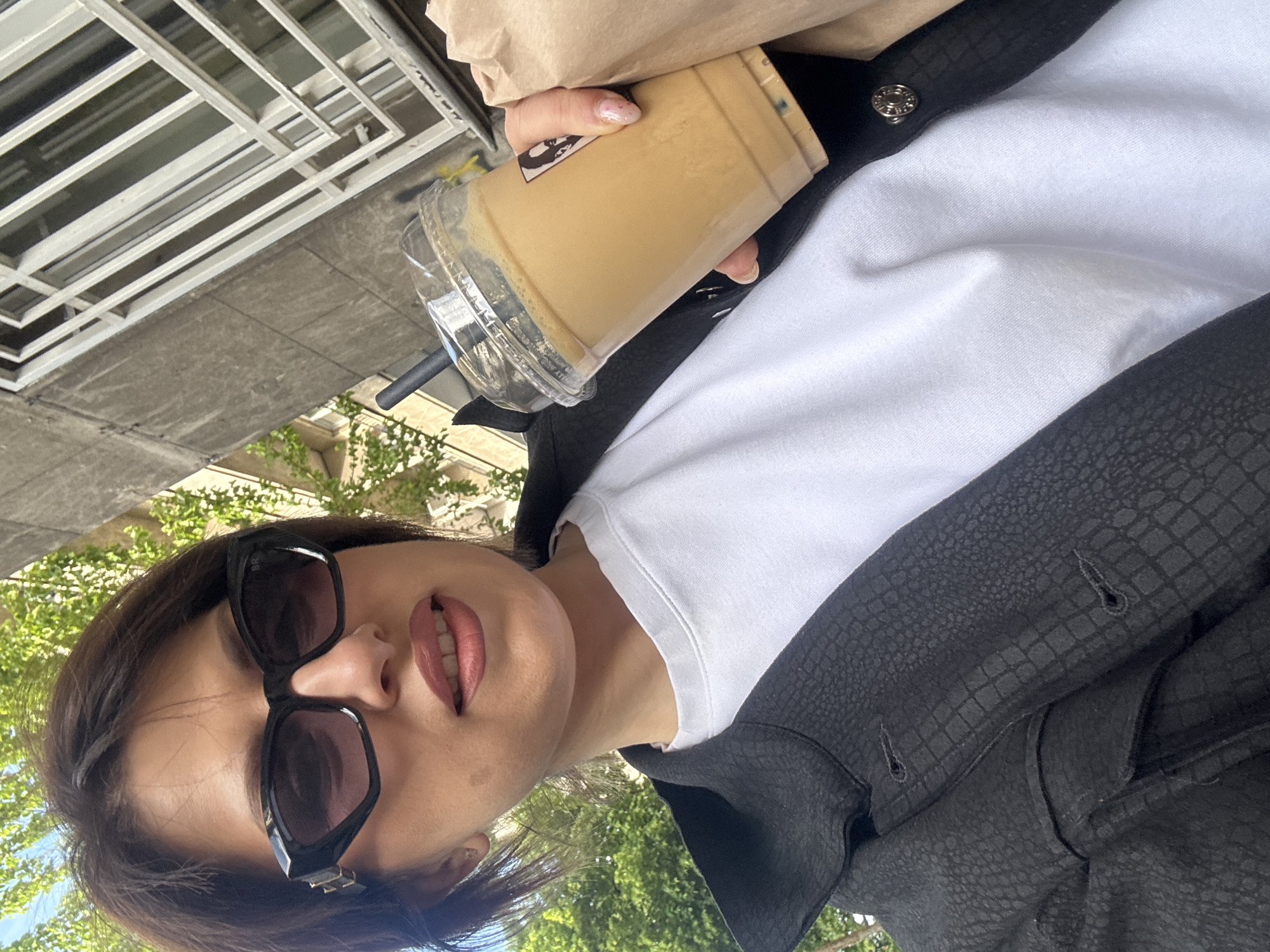 A woman takes a selfie outdoors. She is wearing sunglasses, a white t-shirt, and a black jacket with a textured pattern. She is holding a clear plastic cup with a beige liquid inside. The background includes a building facade, greenery, and a sidewalk. It is plausible that this scene is in Sofia, Bulgaria.