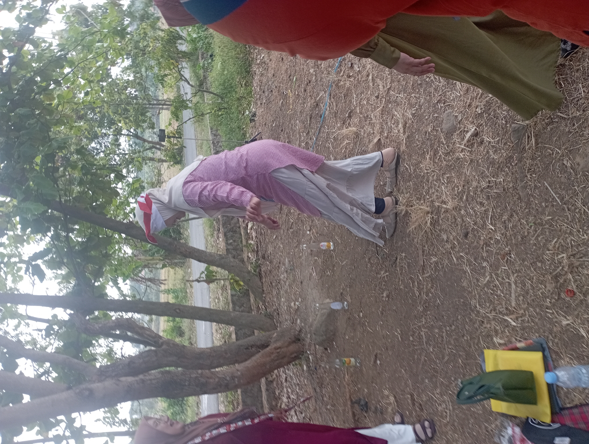The image depicts an outdoor scene, possibly in the city of Purwakarta, Indonesia, where several individuals are engaged in an activity. In the foreground, a person wearing a maroon garment and a headscarf is partially visible on the left. Next to them, a woman in a pink and white striped tunic and a light-colored headscarf with a red and white accessory is in motion, seemingly participating in a game. Her posture suggests she is either about to throw or has just thrown an object. Scattered on the dry, leaf-strewn ground are several empty glass bottles, some appearing to contain liquid. To the right, the back of a person wearing an orange garment and a green dress is visible. Further to the left, a green bag rests on a yellow mat, with a red and black checkered cloth underneath, and a partially visible water bottle. The background features trees with sparse foliage and a road or path beyond, suggesting a rural or semi-rural setting. The overall atmosphere appears casual, with the individuals dressed in modest attire, common in many Indonesian contexts.