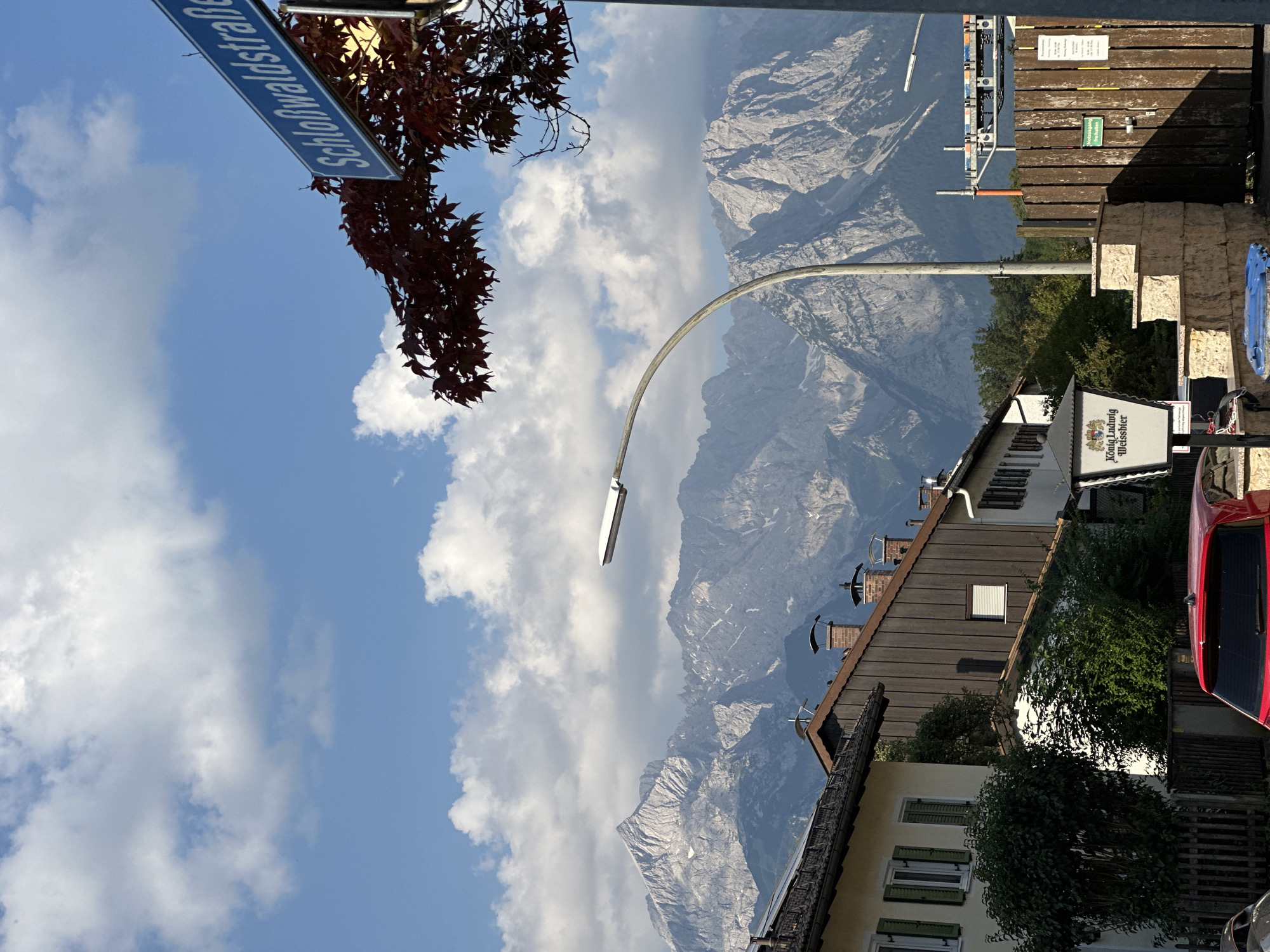 A street sign reading "Schloßwaldstraße" is visible against a blue sky with white clouds. Below the sign, dark red leaves are silhouetted against the sky. In the background, a row of houses with pitched roofs and chimneys can be seen. Behind the houses, a jagged, rocky mountain range dominates the landscape. A modern street lamp curves over the scene. To the right, a wooden fence encloses an area with scaffolding and construction materials. A sign on the fence reads "Notausgang" (Emergency Exit). Further to the right, another sign lists operating hours for a business. In the foreground, the rear of a red car is visible. This scene is located in Garmisch-Partenkirchen, Germany.
