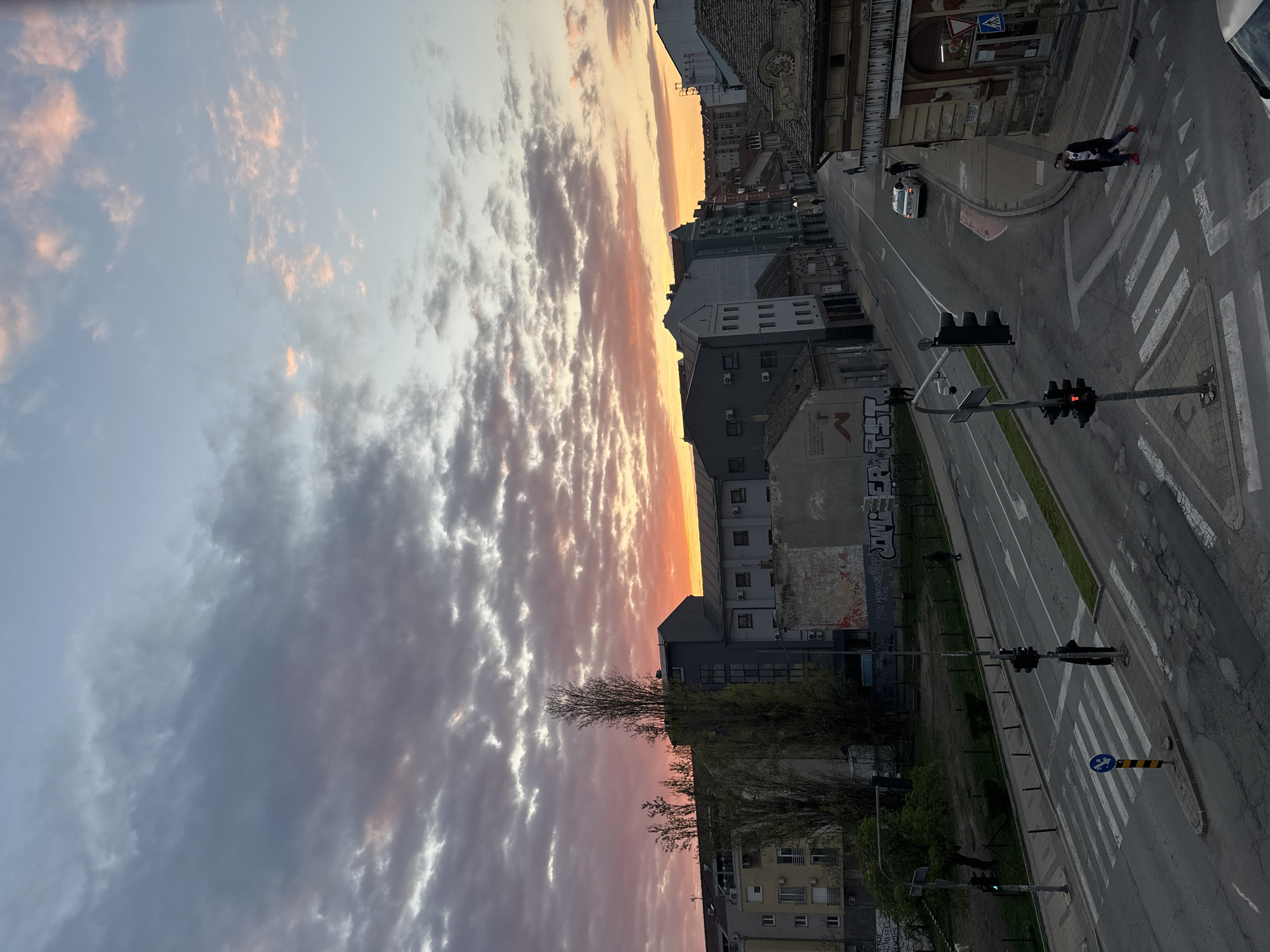 An eye-level, landscape-oriented photograph shows a street in Novi Sad, Serbia, at dusk. The sky transitions from light blue-gray on the left to orange and pink near the horizon, filled with stratocumulus clouds. A road runs from the upper right to the bottom, with sidewalks, buildings, and trees on the sides. A crosswalk with white stripes is visible near the top-right. A pedestrian wearing a dark coat and red shoes is crossing it. A white car is further up the street. Along the road are traffic lights; at least three can be seen, all showing red lights. Graffiti covers some of the buildings along the roadside. The vegetation is green at the bottom, and bare-branched trees are scattered along the road. The overall tone is serene.