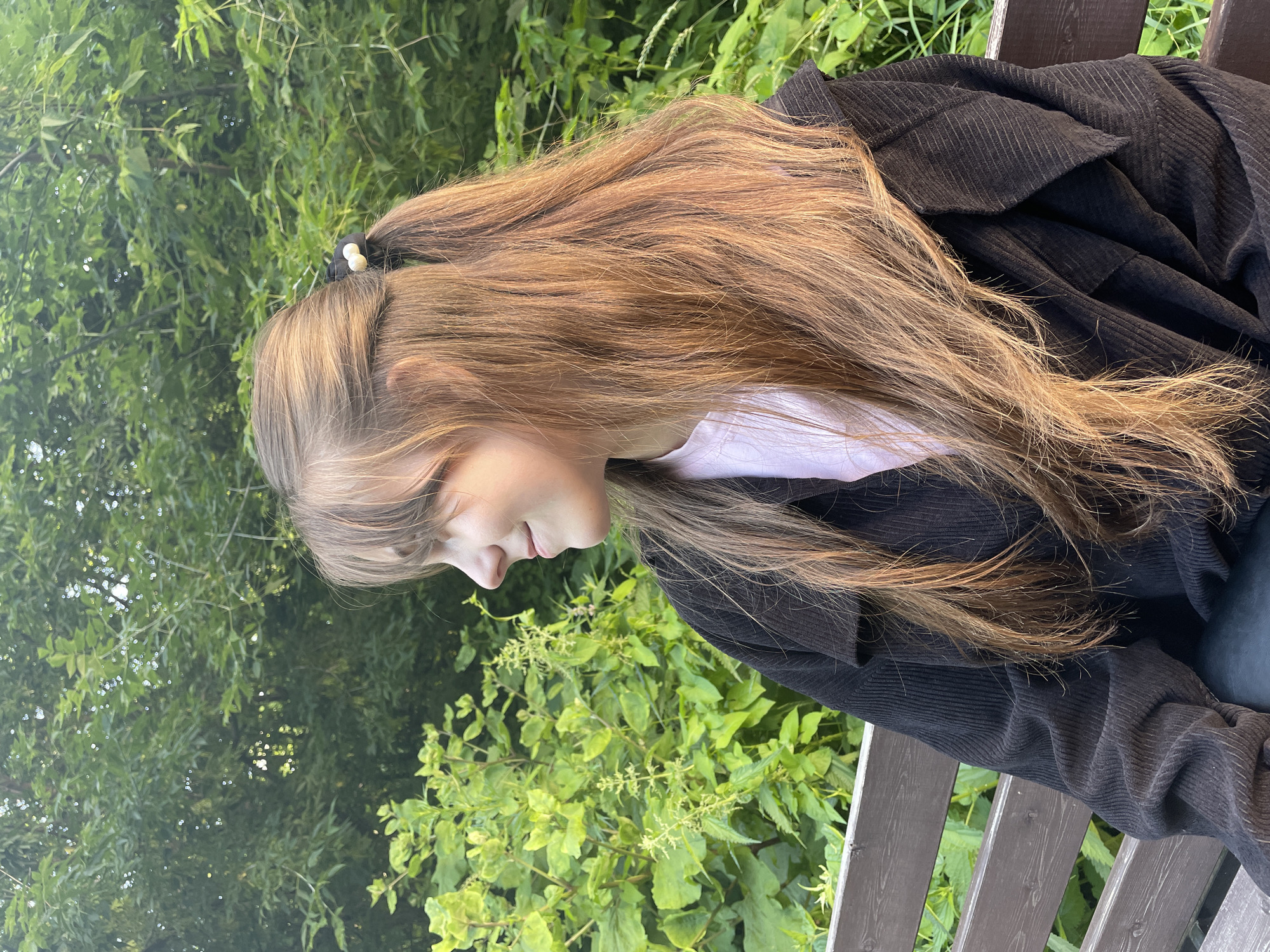 A young woman with long, blonde hair is sitting on a wooden bench outdoors. She is wearing a black corduroy jacket over a white t-shirt. Her hair is pulled back in a ponytail with a small white and black hair accessory. The background consists of lush green foliage and trees, suggesting a natural, outdoor setting. The image is taken from a profile view, showing the woman looking down. The lighting suggests it is daytime, possibly late afternoon, with soft, diffused light. No text is visible in the image. The overall mood appears peaceful and relaxed. The location context provided is Ufa, Russia.