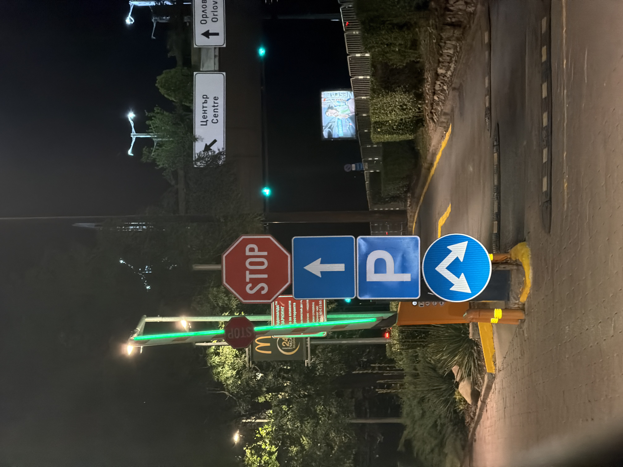 This image captures a night scene in Sofia, Bulgaria, likely near a McDonald's restaurant, indicated by a visible McDonald's sign. The foreground is dominated by a series of traffic signs. Starting from the top, there is a red octagonal "STOP" sign, followed by a blue square sign with a white upward-pointing arrow. Below that is another blue square sign with a white "P" indicating parking. At the bottom of the sign stack is a blue circular sign with two white arrows pointing in opposite diagonal directions, suggesting a lane split. To the left, a green illuminated pole is visible.
In the background, there are directional signs in both Bulgarian and English. One sign indicates "Център Centre" with an arrow pointing left, and another points to "Орлов Orlov" with an upward arrow. A dark screen or billboard displays an image of a person in a green garment. Traffic lights with green signals are visible overhead. The environment includes paved walkways, a road with speed bumps, and lush greenery lining the sides. The overall atmosphere is that of an urban, commercial area at night.