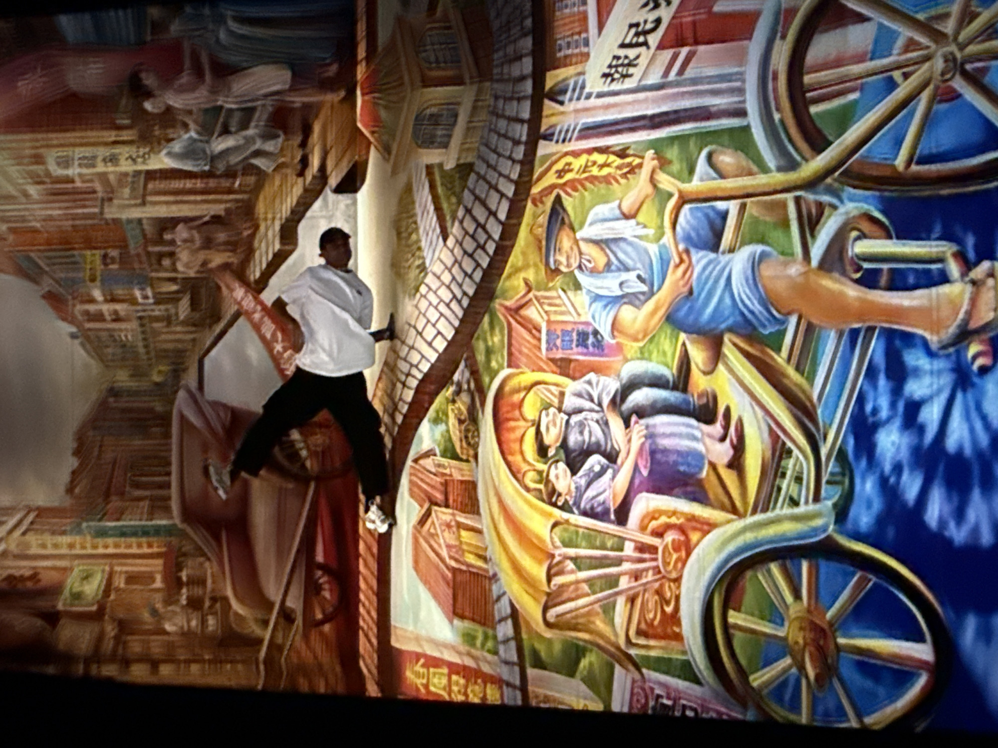 The image depicts a mural painting depicting scenes from a traditional city, possibly in Taipei, Taiwan, given the style of architecture and the presence of a rickshaw and a bicycle with a driver. The mural is divided into horizontal sections, with the top section showing a street scene with buildings, a horse-drawn carriage, and figures walking. A man in white clothing is shown in a dynamic pose, appearing to be breakdancing, with one leg extended and the other bent. The middle section features two traditional-style buildings and a rickshaw with two occupants, a man and a woman, seated under a yellow canopy. The bottom section of the mural shows a man in blue shorts and a light blue shirt, wearing a straw hat, pedaling a bicycle with a rickshaw attachment. He has a towel draped around his neck. The background of this section is a vibrant blue with abstract patterns. Chinese characters are visible on signs throughout the mural, indicating a Chinese cultural context. The overall impression is a vibrant, historical portrayal of urban life.
