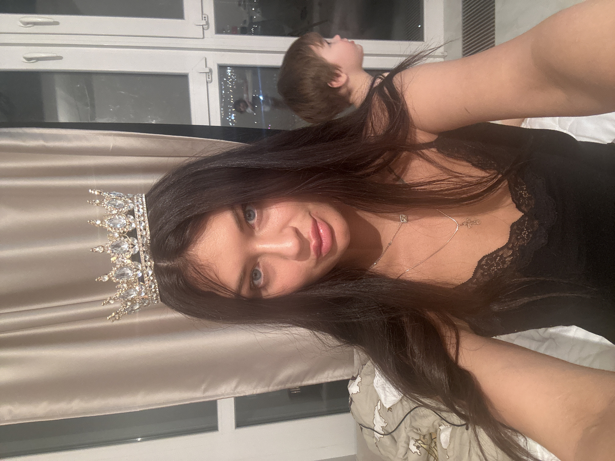 The image captures a young woman and a child in an indoor setting at night, likely a bedroom in Moscow, Russia. The main subject, a woman, is positioned prominently in the foreground, lying down and looking directly at the camera with a subtle smile. She has long, dark hair, blue eyes, and full lips. Adorning her head is a sparkling, ornate crown or tiara encrusted with numerous clear crystals. She is wearing a black top with delicate lace detailing at the neckline and two necklaces: one with a small, square pendant and another featuring a cross pendant.
In the background, slightly out of focus and positioned behind the woman's head and hair, a child with short brown hair is also visible, lying down and looking upwards towards the top right. The indoor setting features white window frames, through which distant city lights are visible against the dark night sky, indicating an urban environment at night. A light brown, silky curtain or blind is partially drawn near the window. White bedding and a patterned duvet are also visible, suggesting a bed. The overall scene appears to be a casual, intimate moment captured at home. There is no visible text in the image.