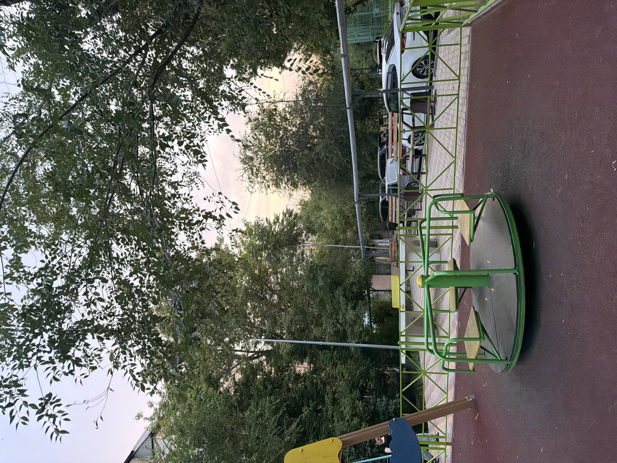 The image depicts an outdoor scene at a playground. A green metal merry-go-round with yellow seating is the central object in the foreground, positioned on a dark red rubberized surface, typical of playground flooring. Behind the merry-go-round, a green metal fence encloses the playground area. Beyond the fence, several white cars are parked in a row, suggesting the playground is adjacent to a parking area or a residential complex.
The background is dominated by lush green trees, their leaves dense and varied in shape, indicating a warm season. The sky is visible through the tree canopy, appearing overcast or hazy, with a soft light suggesting either early morning or late afternoon. The overall atmosphere is peaceful and serene, with no people visible.
To the left of the frame, a portion of a building with a peaked roof is visible, hinting at the surrounding urban or suburban environment. The image is oriented vertically, with the ground and playground equipment appearing to be on the right side of the frame and the trees and sky on the left. The location is identified as Shymkent, Kazakhstan, providing geographical context. There is no visible text in the image.