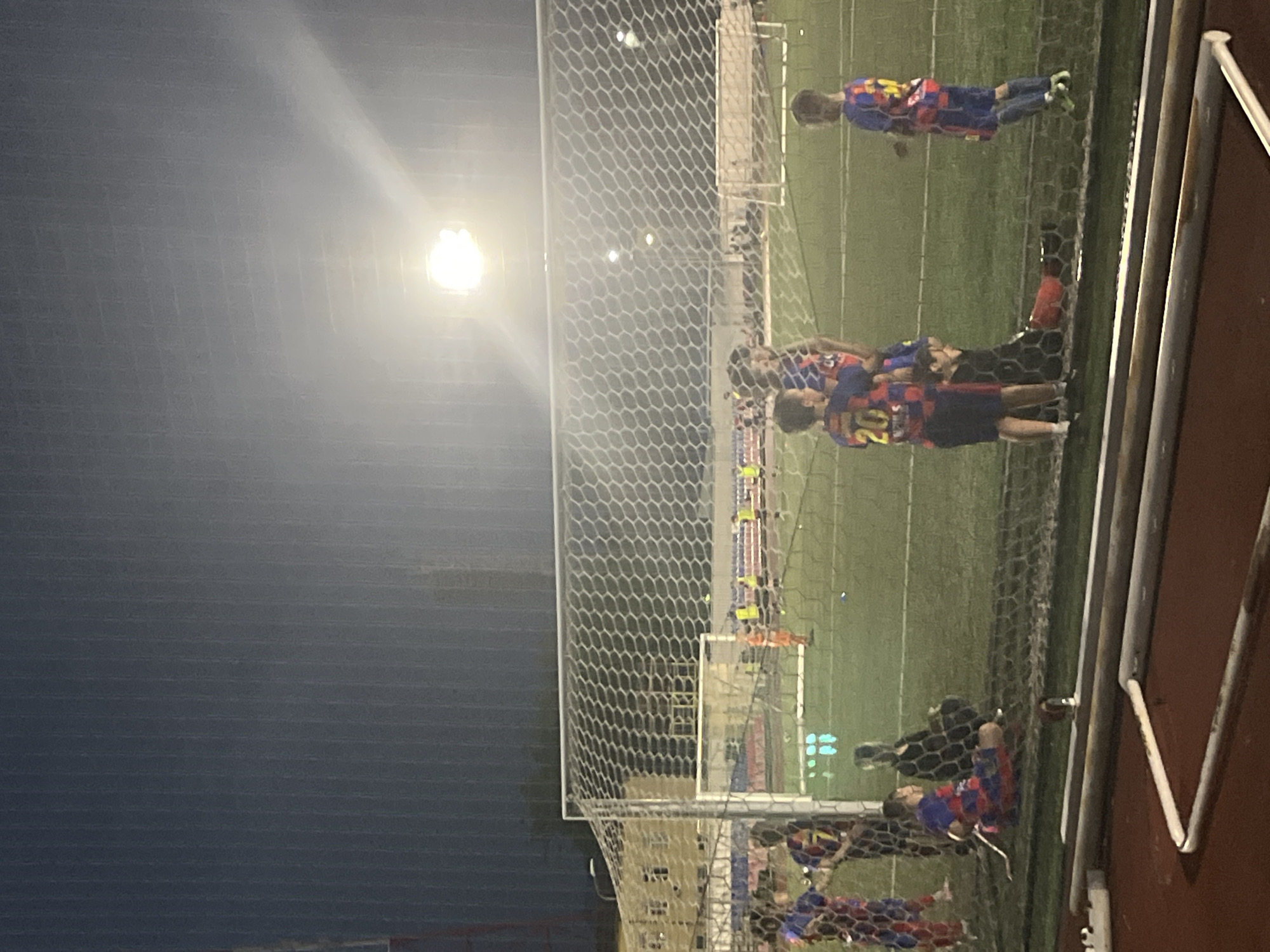 A group of young male soccer players in red and blue checkered uniforms are visible on a green soccer field at night. The field is lit by bright floodlights. In the foreground, a soccer net is partially visible. Some players are standing, while others are sitting or kneeling. One player in a blue jersey with the number 20 is looking away from the camera. Another player in a similar uniform is partially obscured by the net. In the background, other players are scattered across the field, and a goal is visible. The scene appears to be a training session or a game. The presence of floodlights suggests it's an outdoor facility. The architectural style of the buildings in the far background is not clearly discernible due to the low light conditions. The location is identified as the FC Krasnodar Academy.