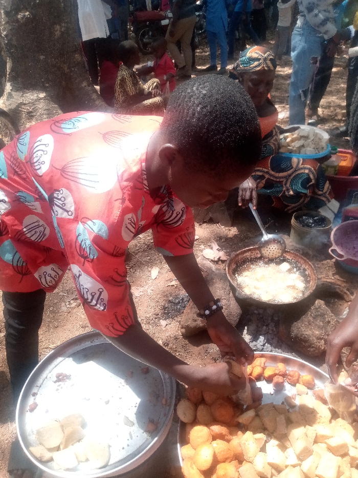 In Kagoma, Nigeria, an outdoor scene depicts food preparation. A young individual in a red, patterned short-sleeved shirt and dark pants is positioned centrally, actively sorting food items on large, round metal trays resting on the bare earth ground. Their hands are engaged with pieces of golden-brown, roundish fried items and lighter, irregularly shaped fried items on one tray. Another tray to the left holds lighter-colored, block-like food pieces.
Behind this individual, an adult female with a patterned headscarf (gele) and dress is seen crouched, utilizing a metal slotted spoon to manage food frying in hot oil within a dark, round pot supported by rocks over a heat source on the ground. A blue plastic bucket containing white food items is visible near her. Multiple other individuals, adults and at least one child in a red shirt, are present in the background, some standing or seated, observing the activity. A tree trunk is visible on the left, and the ground consists of dry earth with scattered leaves. The lighting indicates a bright, sunny daytime environment.