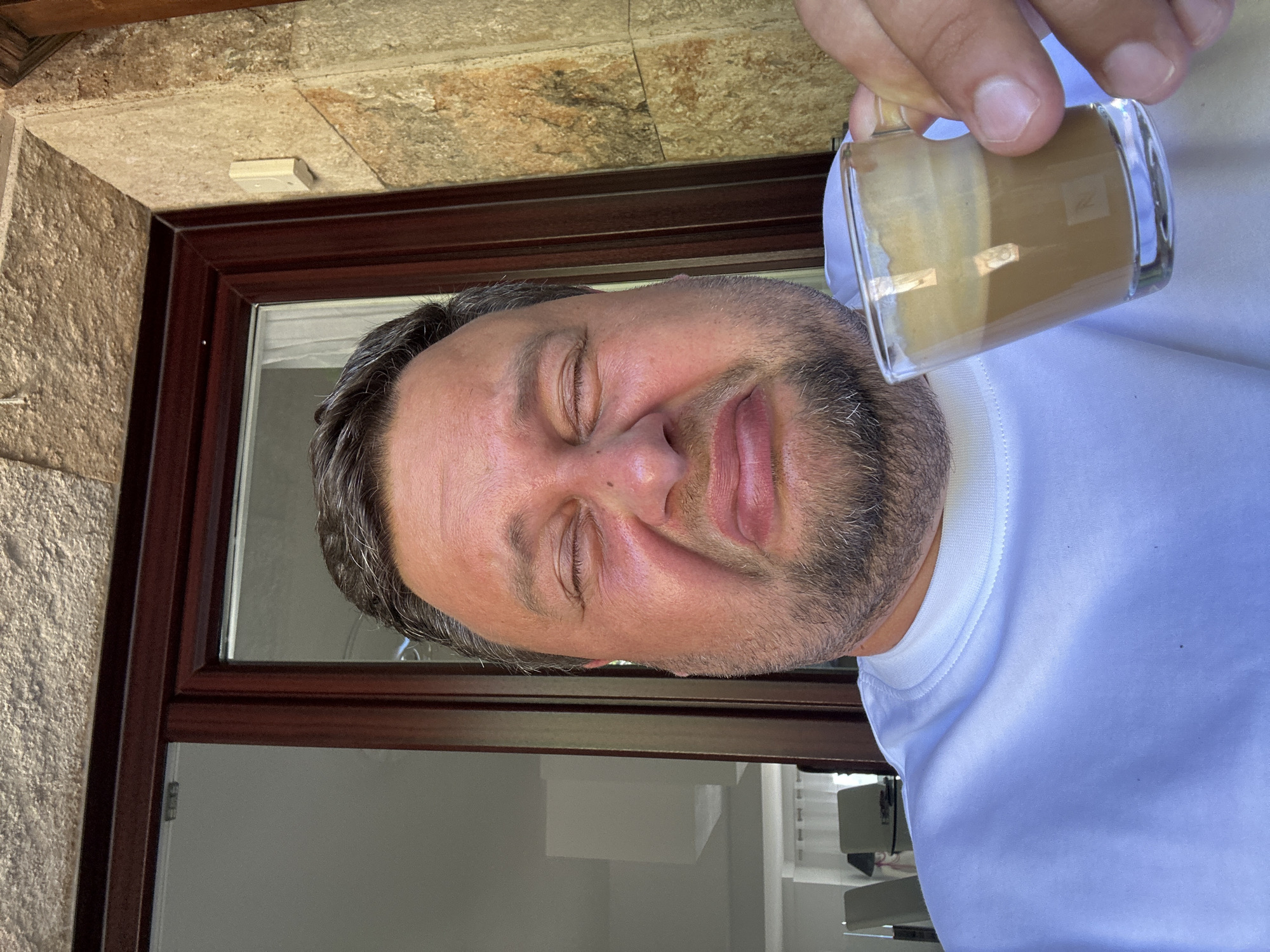 A man with closed eyes is holding a glass of coffee at an angle in front of a brown-framed window. The window is part of a building with stone siding. The man is wearing a white shirt. Based on the architecture and context, the location is potentially Sant Feliu de Guíxols, Spain.