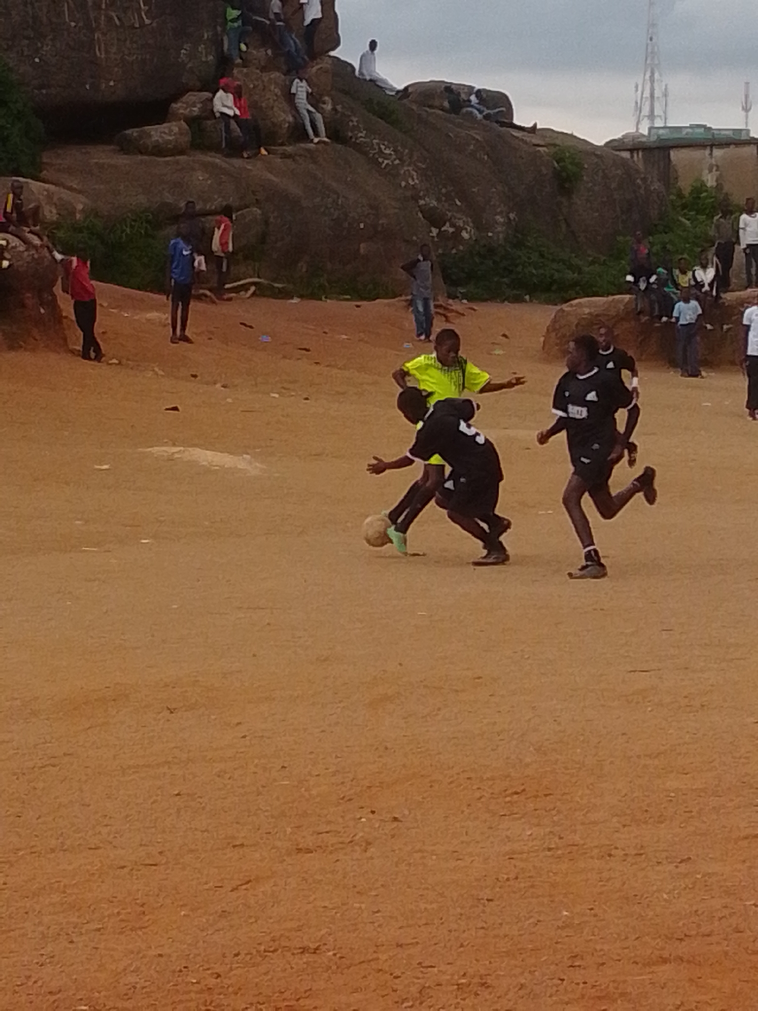 The image depicts an outdoor scene at the base of a large rock formation in Jos, Nigeria. In the foreground, several young boys are engaged in a game of soccer on a dirt field. Three players are in action: one in a neon yellow shirt, and two in black soccer kits. One player in black is dribbling the ball, while another in black is running towards the ball, and the player in yellow is reaching out as if to intercept. In the middle ground, more people are scattered around the base of the rocks, some sitting and observing. The background features the expansive rock formations characteristic of Jos, with some vegetation growing on and around them. A few buildings and a telecommunications tower are visible in the distance, suggesting a semi-urban or peri-urban setting. The overall atmosphere is one of casual recreation within a natural landscape.