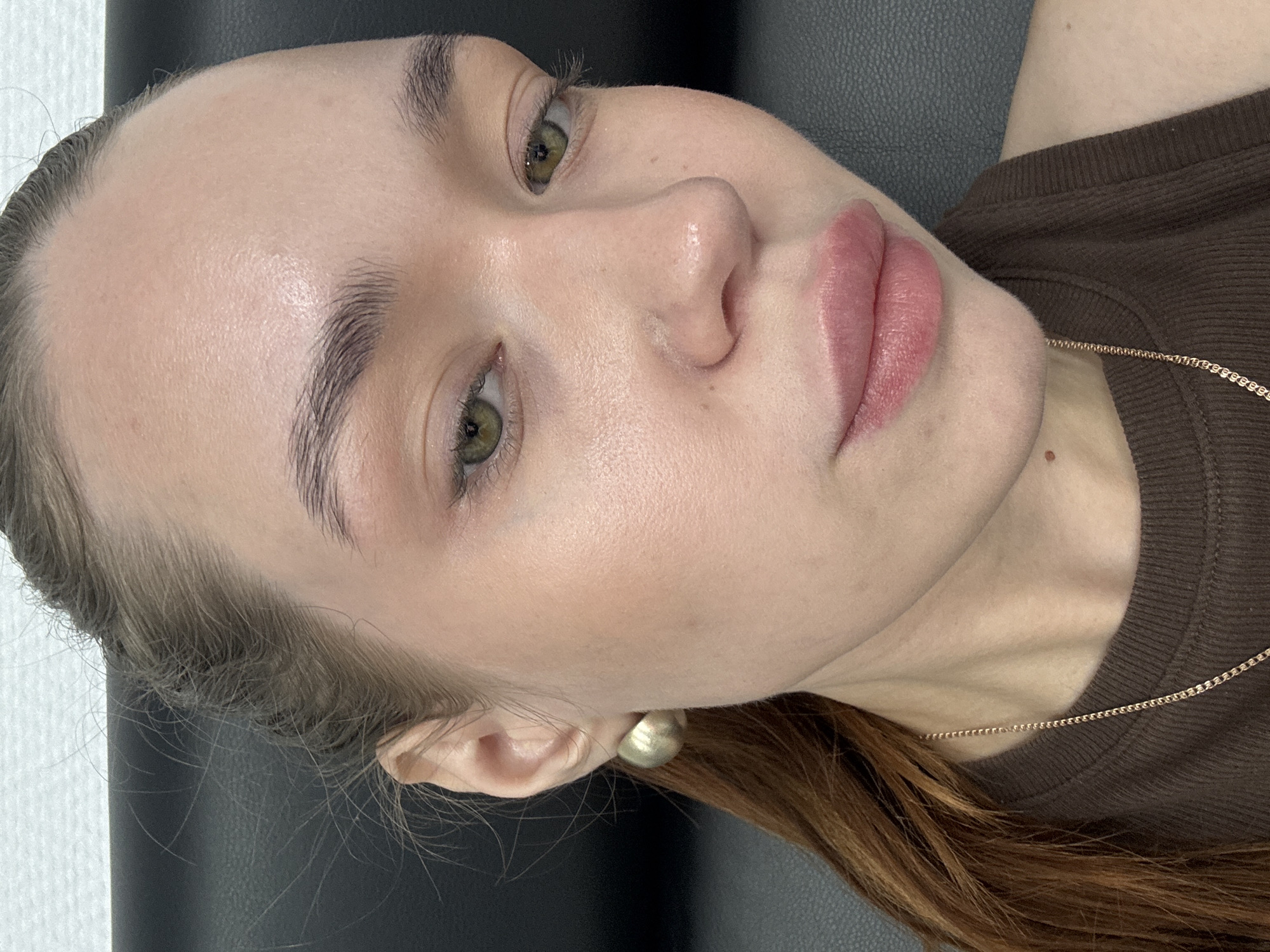 A close-up photograph shows a fair-skinned woman with light brown hair lying on what appears to be a dark leather surface. Her green eyes are open, and she has full, pink lips. She is wearing a brown, possibly knitted, shirt, and a gold chain necklace is visible. A gold stud earring is in her left ear. No specific details in the image indicate the location is Chaikovsky, Russia.