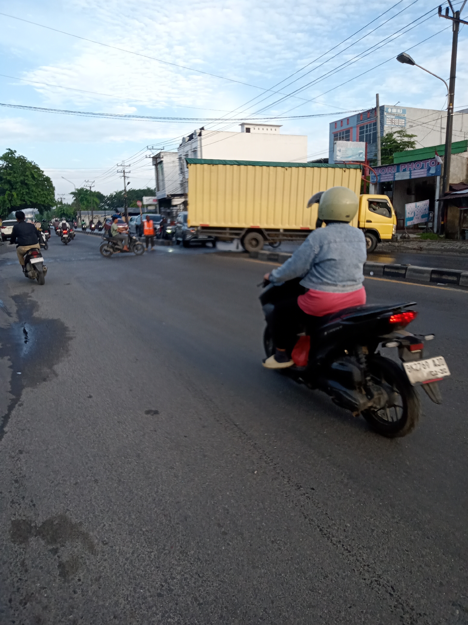 This is a street-level photograph taken in Puji Mulyo, Indonesia, depicting a busy road with vehicular traffic. In the foreground, a person wearing a light blue denim jacket, pink pants, and a light-colored helmet is riding a black motorcycle. The motorcycle has a license plate that reads "BK 2769 AXZ". The rider is looking forward, and the motorcycle's brake lights are illuminated. Behind this motorcycle, a yellow truck with a large, light yellow container is moving. To the left of the truck, other motorcycles and a white car are visible in traffic. Further down the road, more motorcycles are seen, with some riders wearing dark clothing and helmets. On the right side of the road, buildings are present, including one with a sign that says "HOPI PHOTO". The sky is partly cloudy with a light blue hue, and utility lines are strung across the top of the frame. The road surface appears to be asphalt with some dark patches, possibly water or oil stains. The overall scene suggests a typical day with active traffic on a city street.