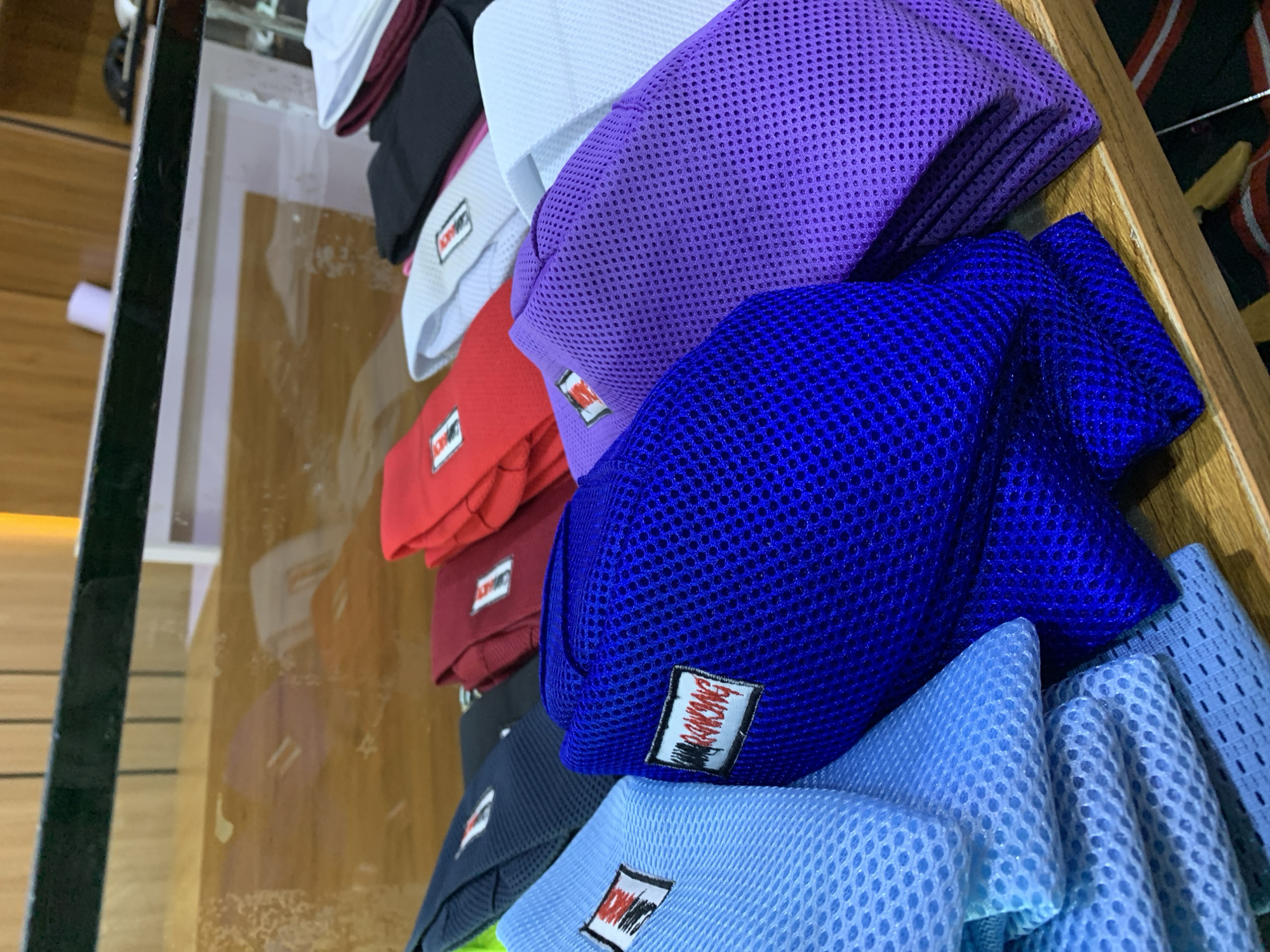 A close-up, eye-level shot displays a stack of folded, mesh-like fabric items, possibly hats or caps, arranged on a wooden shelf in what appears to be a retail setting. The items are varied in color, including light blue, dark blue, royal blue, purple, red, burgundy, black, white, and light gray. Each item has a white rectangular tag with a logo that includes red and black lettering. The background features a wooden wall and a glass display case reflecting the items. The overall lighting is warm and slightly diffused. The location is identified as Gura Top, Nigeria.