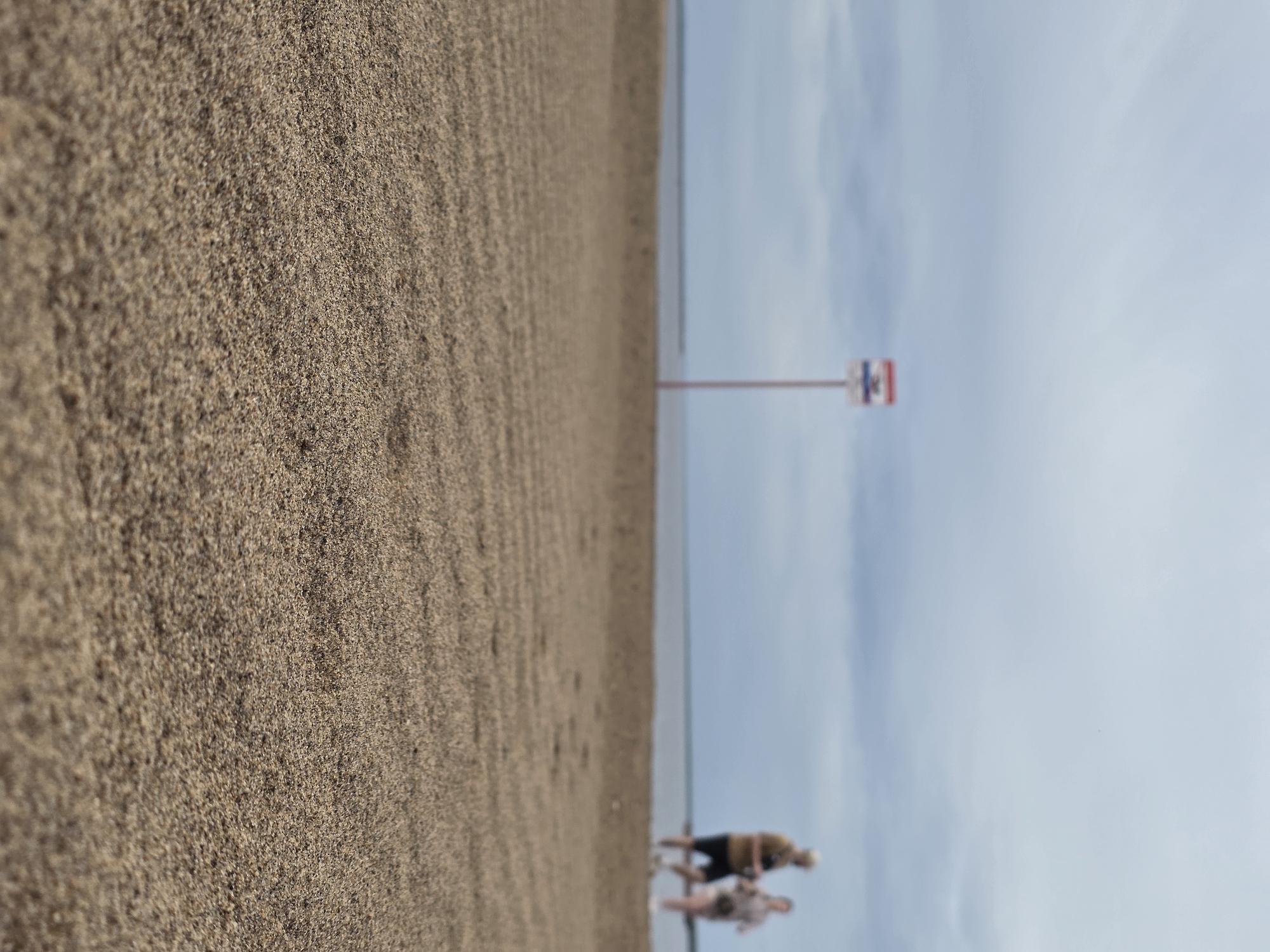 The image shows a beach on the bank of the Kama River in Chaikovsky, Russia. In the background, two people walk by the water. A sign with the city's emblem is visible. The foreground is dominated by sand.