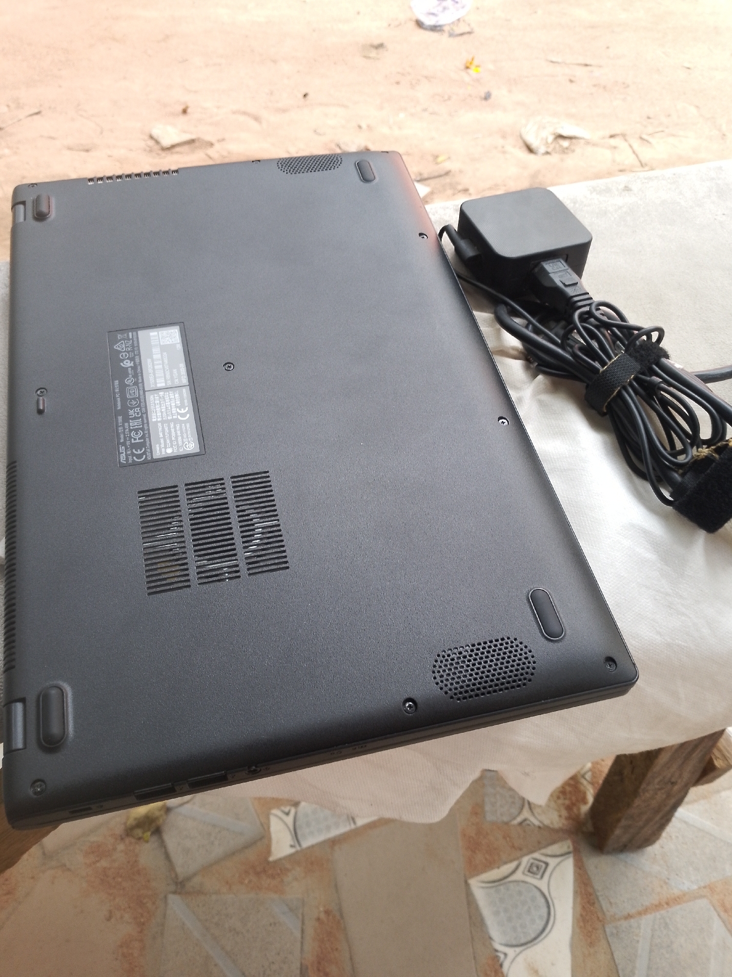 The image shows the underside of a black laptop, lying on a white cloth that is spread over a surface of patterned tiles. Next to the laptop, to the right, is a black laptop charger with its cord bundled and secured with a velcro strap. The laptop has several labels adhered to its base, including regulatory markings like CE and FCC. There are also vents visible on the underside of the laptop. The background is slightly out of focus and appears to be an outdoor setting with sandy ground and some debris. The lighting suggests it is daytime. The scene is located in Jalingo, Nigeria.