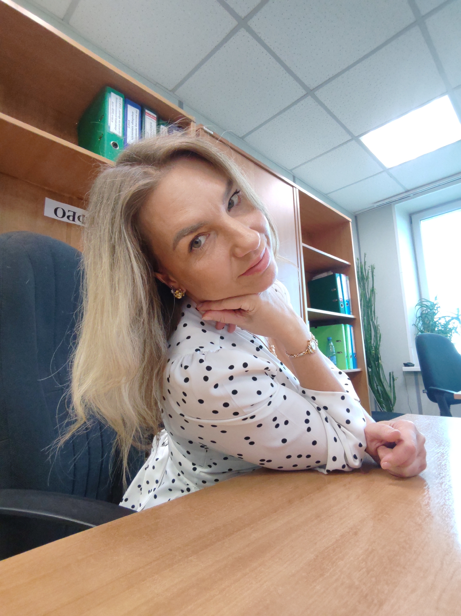 A woman with long blonde hair, light-colored eyes, and fair skin is captured in a medium shot, looking up towards the camera with a gentle smile. She is wearing a white blouse adorned with black polka dots and has her right hand gently resting under her chin, while her left arm rests on a wooden desk. Gold-colored earrings and a matching wrist watch/bracelet are visible accessories.
The setting appears to be an office environment. The woman is seated at a dark office chair in front of a light-colored wooden desk. Behind her, a wooden shelving unit holds several binders in green and blue. A long, green, succulent-like plant stands on the floor next to the shelving unit, near a window that shows a bright outdoor light, though no specific weather conditions are discernible. The ceiling features white acoustic tiles with integrated fluorescent lighting. Another dark office chair is partially visible near the window.
Visible text includes a label "OAD" (likely Cyrillic "ОАО") affixed to the bottom shelf. On the binders, some Cyrillic text can be partially deciphered. On the top shelf, a green binder has text that seems to indicate "Закупки 2020 год" (Purchases 2020 year). A blue binder next to it reads "АКТЫ" (Acts) followed by text possibly related to "внутренний" (internal). On the lower shelf, a green binder clearly displays "Бухгалтерия" (Accounting) and "ООО" (Limited Liability Company). The overall impression is that of a professional and organized workspace.