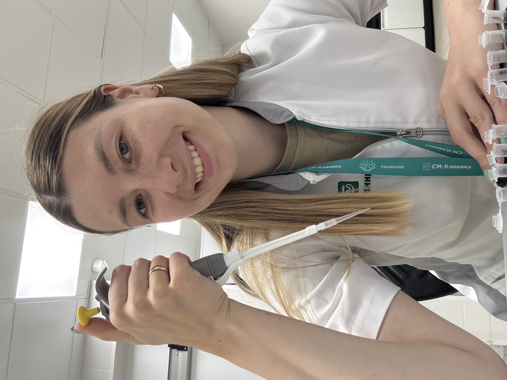 The image is a rotated selfie, depicting a young woman in a laboratory or medical setting, smiling brightly at the camera. The photograph is oriented horizontally, with the subject's head on the left side of the frame.
**Subjects:** The primary subject is a young woman with long, straight, light brown hair, light-colored eyes, and freckles. She is wearing a white lab coat over a light brown or khaki top. A gold hoop earring is visible in her right ear, and a simple gold band ring is on her left ring finger. Her nails are painted with a light green polish. Her left hand holds a grey and white micropipette with a yellow ejector button, pointed towards the right. Her right hand is visible on the far right, reaching towards or holding a rack of small, clear plastic tubes, suggesting laboratory work.
**Scene and Setting:** The background is an indoor, well-lit environment with white walls and what appears to be a white grid ceiling. A bright light source, possibly a window, is visible in the upper left background. The overall setting strongly suggests a clinic or scientific laboratory.
**Activity:** The woman is actively engaged in a laboratory task, holding a micropipette, implying she is preparing samples or performing an assay. Her broad, joyful smile indicates a positive and pleasant demeanor while working, possibly taking a break to capture the moment.
**Notable Details:** The woman’s expression is one of happiness and approachability. Around her neck, she wears a teal-colored lanyard with white text and logos. Visible text on the lanyard includes "СМ-Клиника" (SM-Klinika), which appears multiple times, along with "УВАЖЕНИЕ" (UVAZHENIE, meaning "RESPECT") accompanied by a handshake icon, and an Instagram-like logo also next to "СМ-Клиника." Given the context of Moscow, Russia, "SM-Klinika" refers to a known network of medical centers in Russia.
