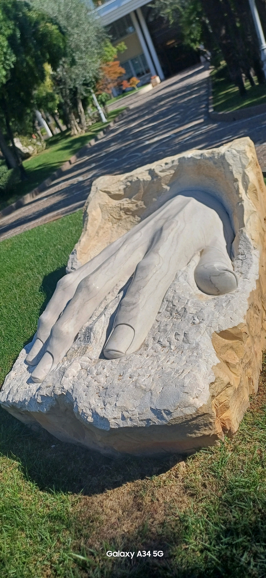 This is a close-up, vertical, outdoor photograph of a stone sculpture of a human hand emerging from a large, rough-hewn rock. The sculpture is rendered in a pale, marble-like material and exhibits significant detail in the fingers, nails, and the creases of the skin. The rock itself is a rough, beige-colored sedimentary stone with a textured surface. The sculpture is positioned on a patch of grass, with some areas of dry, brown grass interspersed with vibrant green. In the background, out of focus, are trees, a paved walkway with a pattern of shadows cast by the trees, and a modern building with a light-colored facade and large windows. The lighting suggests it is daytime, with strong sunlight creating sharp shadows. The bottom right corner of the image displays the text "Galaxy A34 5G". The scene appears to be in a public park or garden setting.