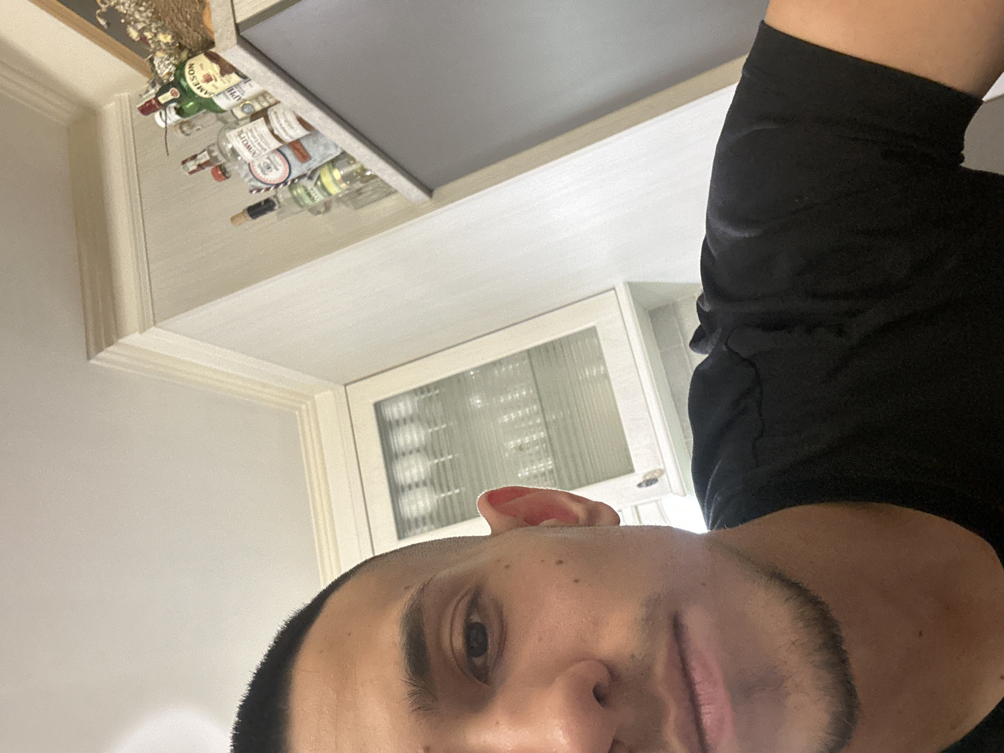 A close-up, low-angle selfie shot captures a male subject indoors in Plovdiv, Bulgaria. The subject, positioned on the lower left and center, is a young adult male with a very short buzz cut, dark eyes, and a neatly trimmed dark beard or goatee along his jawline and chin. He is wearing a plain black t-shirt. The background reveals an interior space, likely a kitchen or dining area, with light-colored walls and a white ceiling featuring crown molding. To the left of the subject, a white cabinet with a glass panel door is visible, showcasing several clear drinking glasses arranged inside. To the upper right, a shelf or countertop holds an assortment of various liquor bottles, including brands like Jameson Irish Whiskey and Dewar's White Label Scotch Whisky, alongside a decorative item composed of dried flowers or foliage. The scene is illuminated by ambient indoor lighting, casting soft shadows. The overall perspective is tilted, with the ceiling line and background elements receding diagonally from left to right.