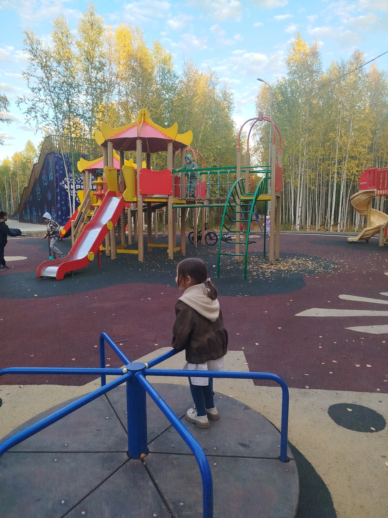 The image depicts a children's playground located in the city of Mirny, Russia. The playground features a large, multi-level play structure with red and yellow accents, including a red slide, green climbing nets, and a yellow spiral slide visible in the background. The ground surface is a dark red and grey rubberized material, with scattered autumn leaves indicating the season.
In the foreground, a young girl with dark hair, wearing a brown jacket and white shoes, is seen from behind standing on a blue merry-go-round. The merry-go-round has a grey circular base divided into sections and a central blue pole with supporting blue bars.
Several other children are present. To the left of the play structure, a child in a grey and white plaid shirt and a white hat is on a scooter. Another child in a dark outfit is standing nearby. In the middle ground, near the play structure, a child in a green hooded outfit is visible on the platform.
The playground is surrounded by a dense forest of birch trees with yellowing leaves, suggesting it is autumn. The sky above is blue with scattered white clouds. The overall scene is that of an active outdoor recreational space for children during daylight hours.