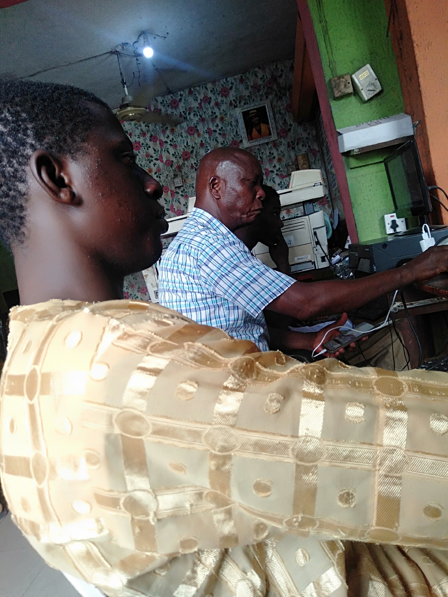 The photograph captures an interior scene in Shangisha, Nigeria. An older man with a bald head and wearing a blue and white checkered shirt is seated at a desk, with hands on a desk. There are multiple printers and electronic devices visible behind him. The foreground is dominated by the shoulder and back of another person wearing a gold-colored fabric with a geometric pattern. A ceiling fan and a single light bulb hang from the ceiling. Wallpaper with a floral pattern can be seen on the walls behind the man, with an electrical box on the right.