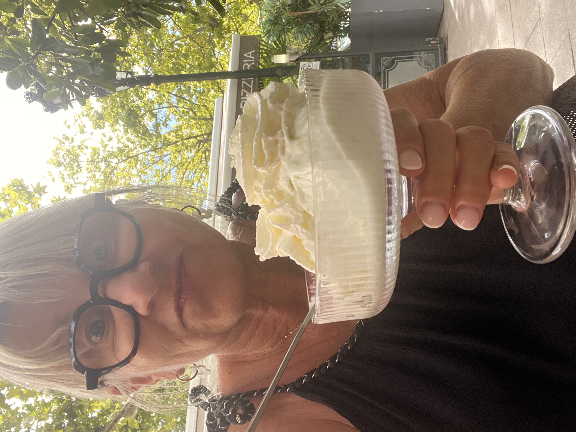 A medium shot captures a woman from the chest up, outdoors, holding a dessert in a clear glass. She is wearing a black tank top and large, dark-rimmed glasses. Her blonde hair is pulled back. The dessert appears to be ice cream or a similar frozen treat topped with a generous swirl of whipped cream. A silver spoon is visible in the dessert.
Behind her, a green awning with the partial word "PIZZERIA" is visible, suggesting the location is a restaurant. A street lamp and lush green trees with dappled sunlight filtering through them indicate an outdoor setting. The overall atmosphere is casual and bright. The woman's facial expression is a close-up, and her eyes are wide, looking directly at the camera. Her skin shows signs of sun exposure, and she is wearing a delicate necklace. She is outdoors in Montélimar, France.
