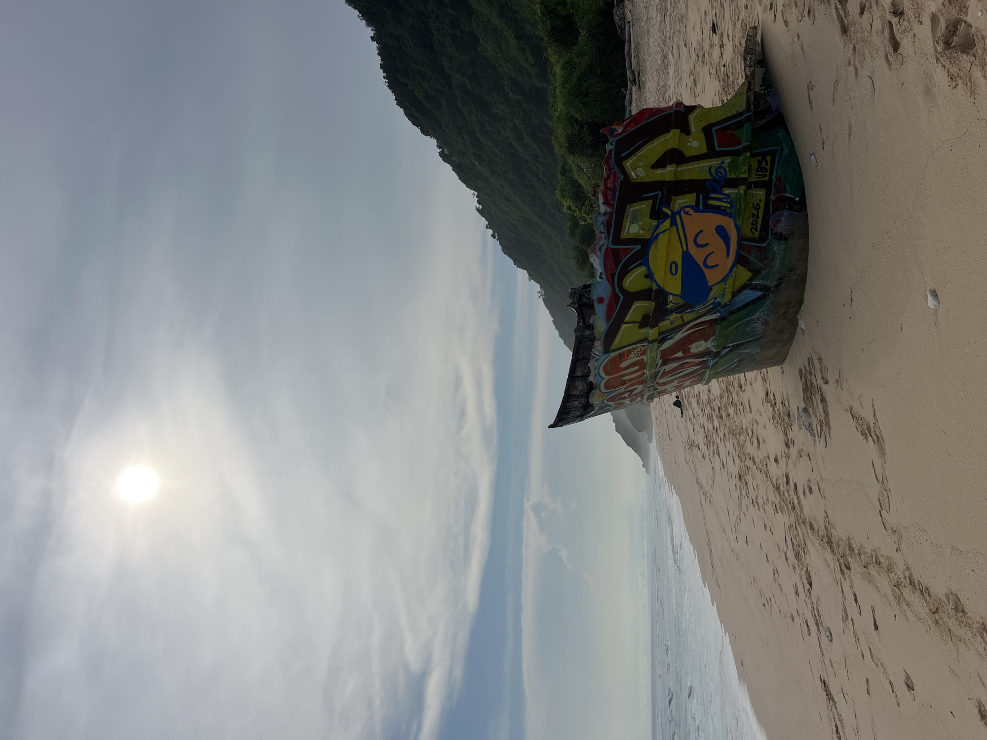 A beached boat sits on the shoreline of a beach in Pecatu, Indonesia. The boat is covered in graffiti, featuring cartoonish designs and lettering. A densely forested cliff rises in the background, indicating the rugged coastal terrain. The beach is sandy, and footprints are visible. The sky is partly cloudy with the sun shining brightly through the haze. The sea is visible in the distance, meeting the sky at the horizon.