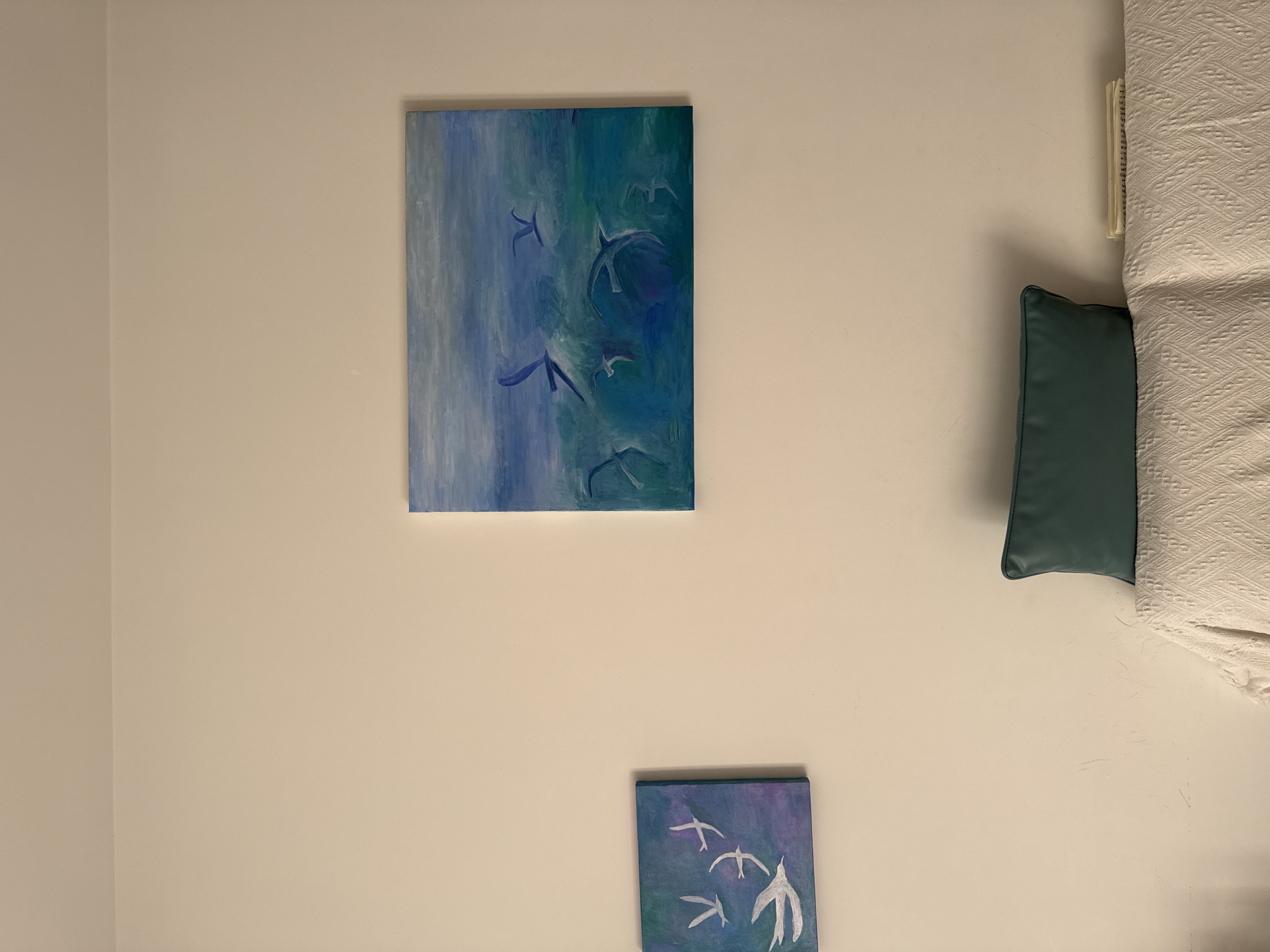 The image shows a wall in a room. On the wall are two paintings. The larger painting is a vertically oriented rectangle with blue, green, and white hues. It depicts several stylized birds. Below and to the right of the larger painting is a smaller, square painting, also depicting stylized birds in flight. A bed with a white blanket and a green pillow is partially visible on the right side of the image. The wall is off-white. There are no people visible in the image. The location is not specified.