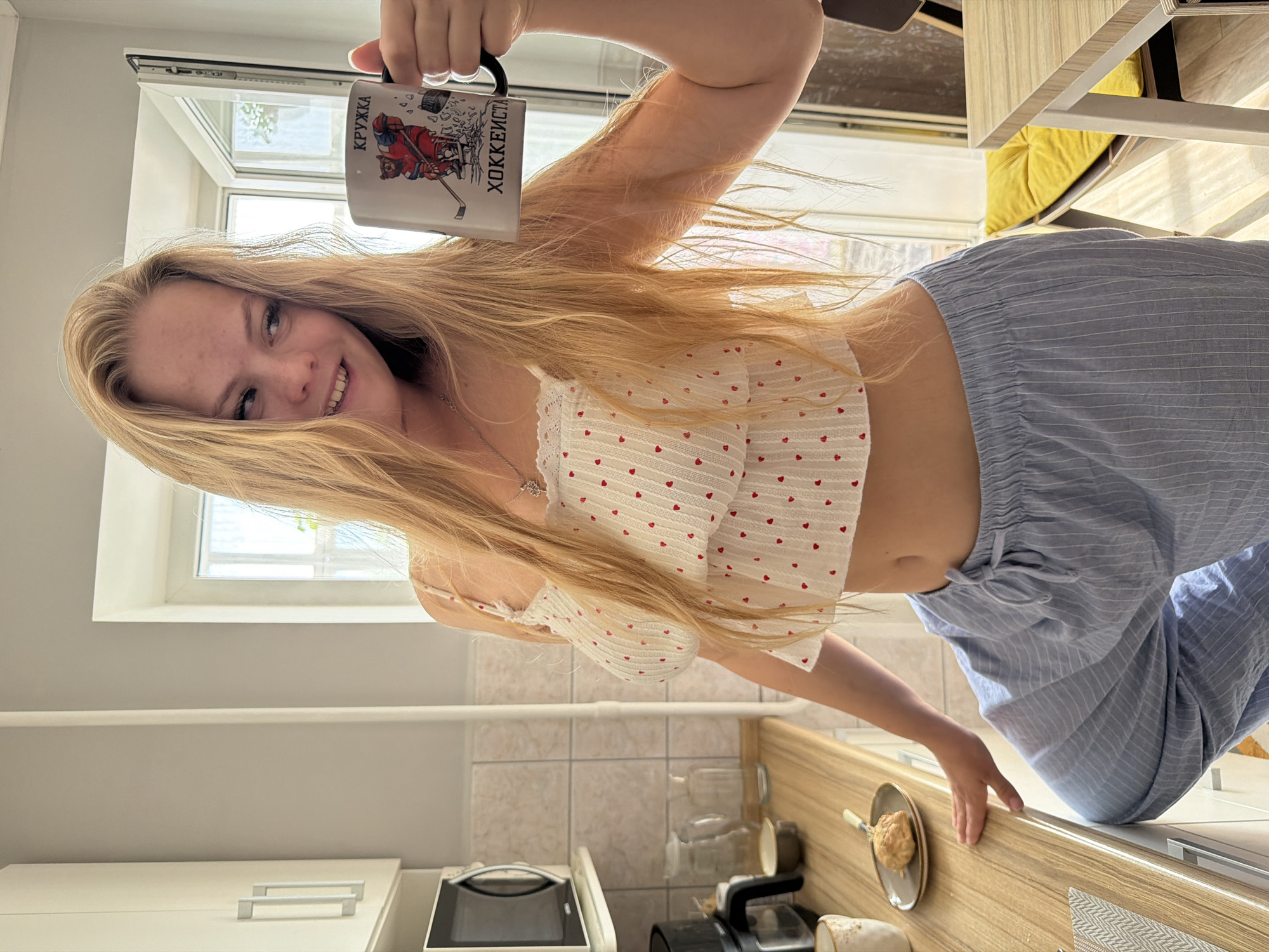 A young blonde woman stands in a sunlit kitchen, holding a mug with a hockey-themed image and Russian text. She is wearing a white, heart-patterned crop top and light blue striped pajama pants. The kitchen features light-colored cabinets, a microwave, and a countertop with various items, including a small plate with a pastry. A window in the background shows a glimpse of the outdoors, suggesting a domestic setting. The presence of the mug with "Chaikovsky" (Чайковский) text, a city in Russia known for its hockey tradition, and the overall domestic environment, points to the location being a kitchen in Chaikovsky, Russia.