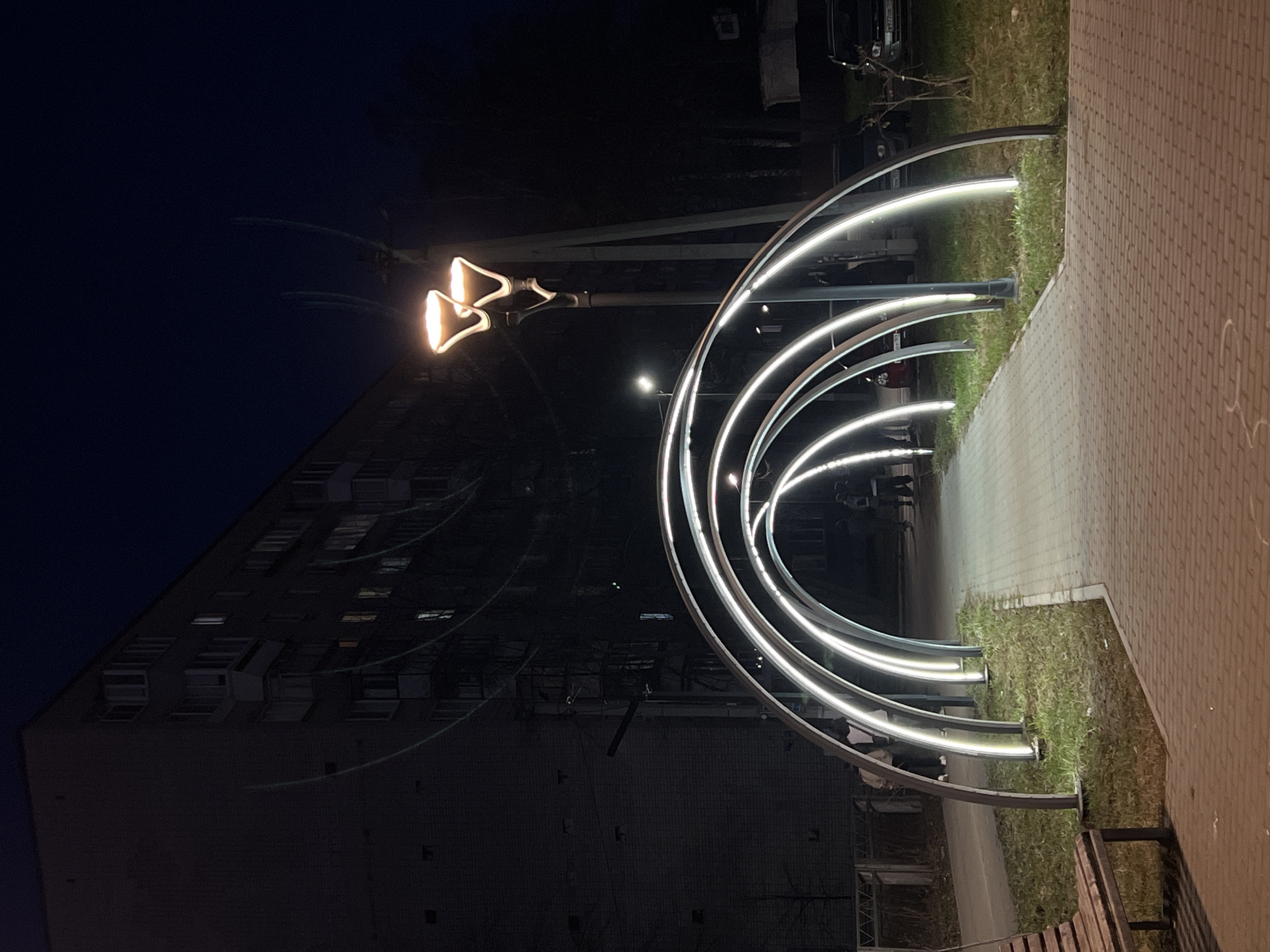 The image, taken at night in Chaikovsky, Russia, depicts a well-lit pedestrian area adorned with modern, illuminated architectural features. The scene, rotated 90 degrees clockwise from its correct orientation, shows a dark night sky.
In the foreground and midground, a series of contemporary arched structures create a glowing pathway. These arches are constructed from dark frames with bright white LED lights tracing their curves, forming a luminous tunnel-like effect. To the right of these arches, a paved pathway, likely made of light-colored bricks or pavers, is visible. A strip of green grass runs parallel to the pathway, separating it from the illuminated arches. Beyond the arches, another grassy area and some bare trees are visible.
A prominent streetlamp with two distinct, upward-facing, horn-shaped lights stands near the center of the frame, casting a warm, yellowish glow. Further down the path, a smaller, standard streetlamp is also visible, contributing to the overall illumination. In the background, a large, dark multi-story residential building stands to the left, with a few windows dimly lit. Several cars are faintly visible parked in the distance, behind the archways and streetlamp.
Faint, blurry figures of at least two people can be seen walking through the illuminated archways, moving away from the viewer, suggesting an evening stroll or commute. One person appears to be wearing a light-colored top. A wooden park bench is partially visible in the bottom right corner of the paved path. The atmosphere is peaceful and inviting, showcasing a modern urban or park environment after dark. No visible text is present in the image.
