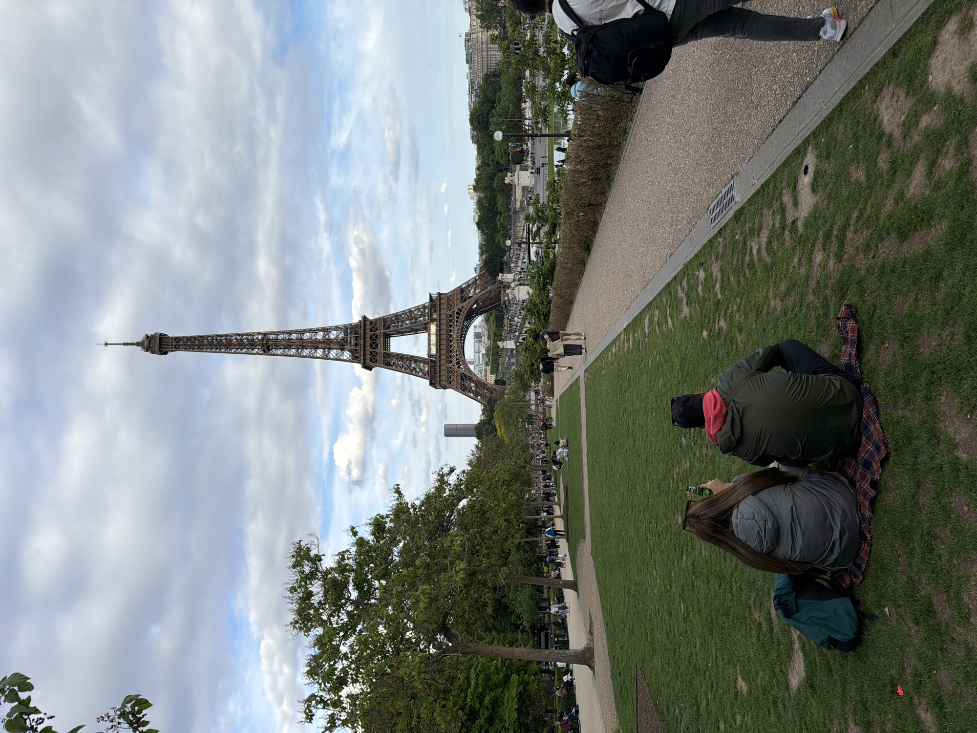 The image captures the Eiffel Tower in Paris, France, from an elevated vantage point within a park setting. The tower is the focal point, standing tall against a partly cloudy sky. A row of trees lines a walkway leading towards the tower, with many people sitting along the walkway and on the grassy areas. In the foreground, two people are seated on a blanket on the grass, appearing relaxed and engaged in leisure. Another individual is partially visible at the top right of the image, walking along a paved pathway. The environment is characterized by green grass, trees, and the cityscape in the background, presenting a scene of leisure and tourism in Paris.