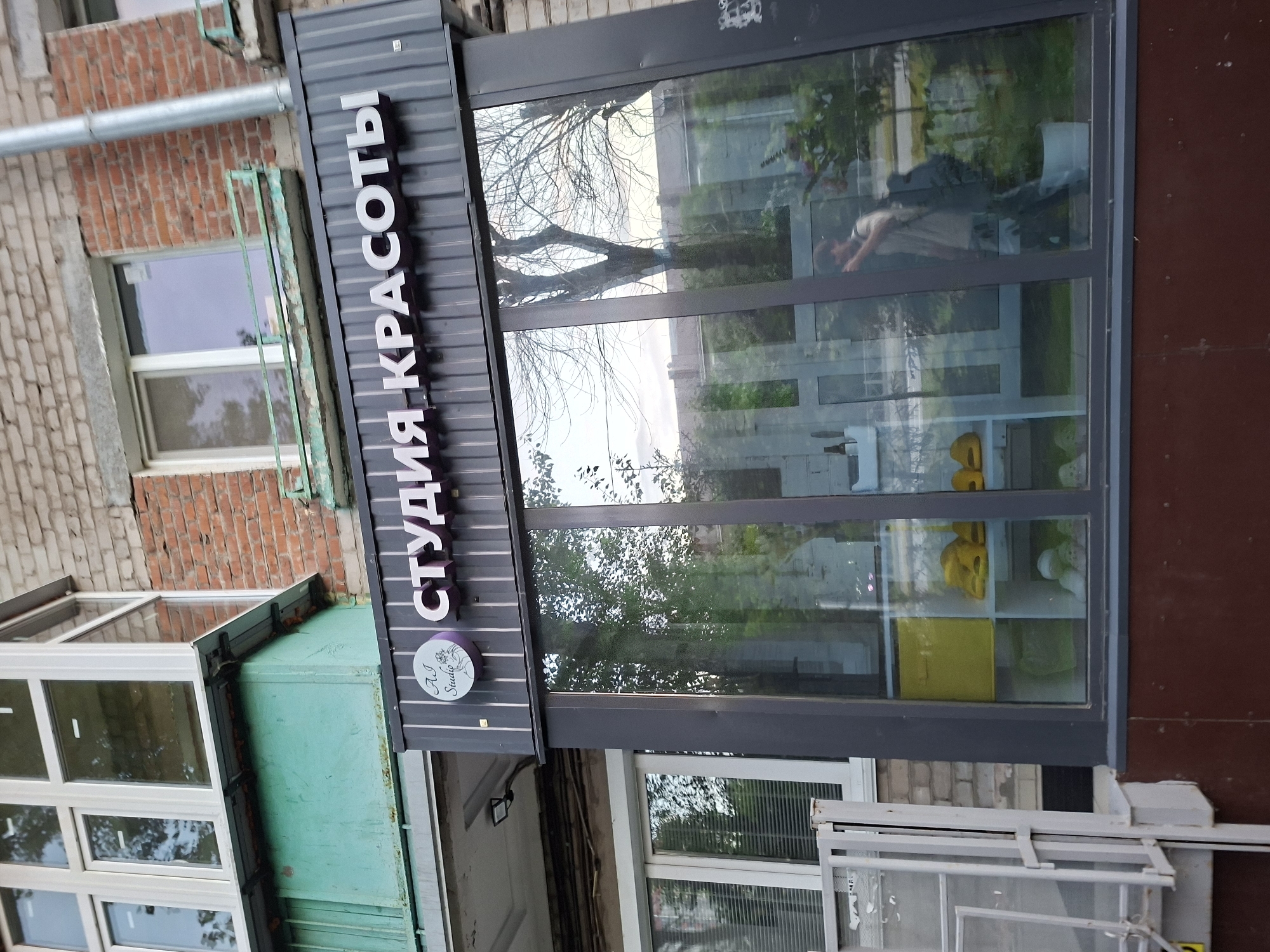 This image shows the storefront of a beauty salon named "СТУДИЯ КРАСОТЫ" (Beauty Studio) located in Kazan, Russia. The salon features a dark gray facade with large glass windows reflecting the surrounding environment. The windows display interior shelving with yellow and white items, possibly products or decorative elements. A person wearing a light-colored t-shirt and backpack is visible in the reflection, walking past the storefront. Above the storefront, a brick building with windows and a green balcony is visible. The overall scene depicts an urban commercial setting.