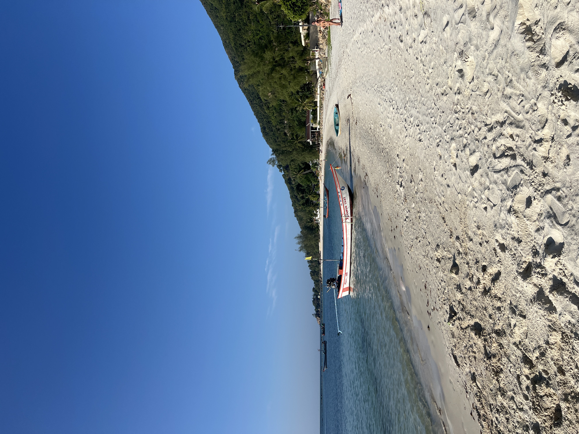 A sunny beach on Ko Pha-ngan District, Thailand, features a white sand shoreline meeting clear blue ocean water. A prominent red and white fishing boat is moored near the shore, with a yellow flag attached to a pole on its stern. Several other boats are visible further out in the water. The background reveals a densely vegetated hillside, characteristic of the island's landscape. Two individuals, one wearing a bikini, stand on the sand. The sand itself is marked with footprints. The sky is a clear, deep blue.