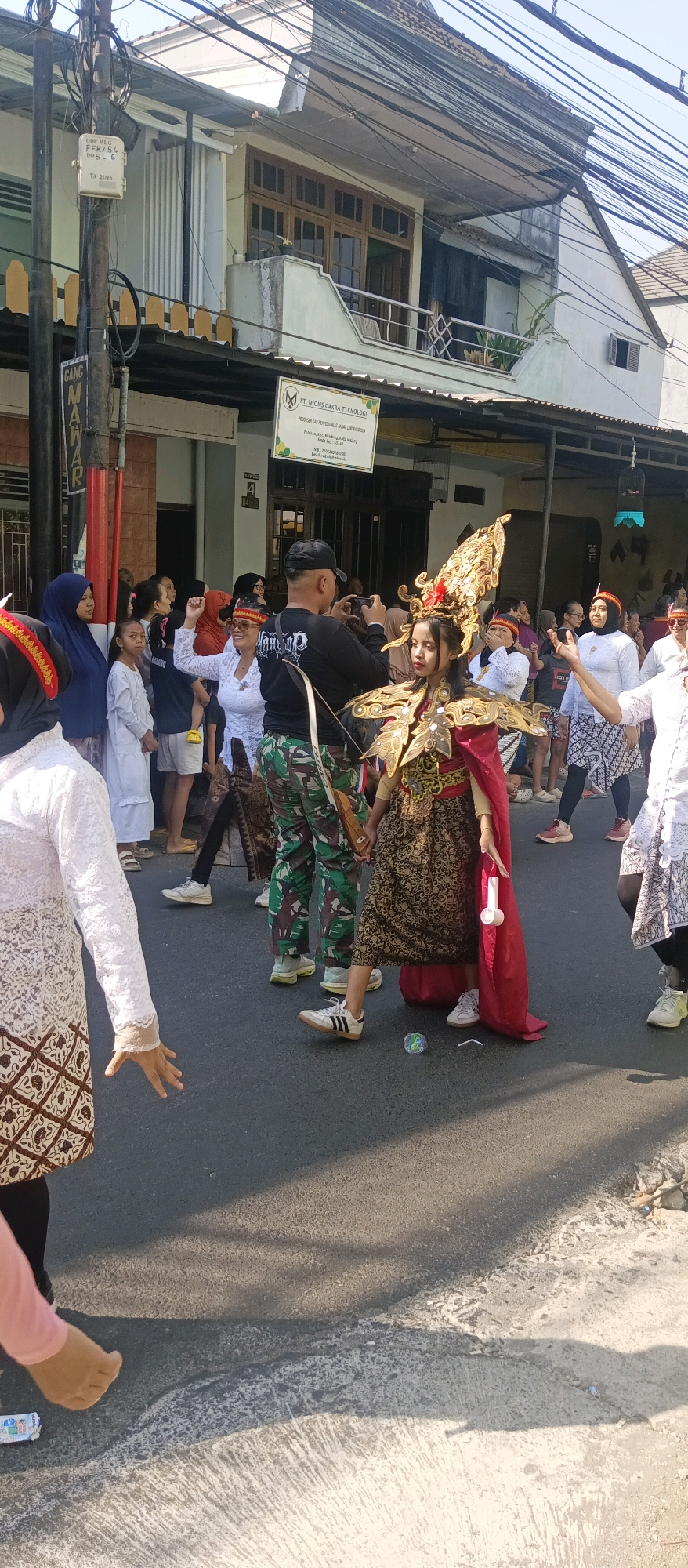 The image depicts an outdoor street scene in Malang, Indonesia, likely during a cultural event or parade. In the foreground, a young girl adorned in an ornate golden headdress and a traditional patterned skirt is the central focus. She is holding a bow and arrow, suggesting a ceremonial or performance role. To her left, a man in a black t-shirt and camouflage pants is seen taking a photo with his phone.
Behind them, a crowd of people of various ages are gathered, observing the event. Many are dressed in traditional Indonesian attire, including headscarves and batik patterns. A woman in a white lace outfit with a red headband is enthusiastically raising her arms. The background shows a street lined with buildings, some appearing to be shops or residences, with visible power lines and street signs. One sign reads "PT. MIONS CAKRA TEKNOLOGI", indicating a business establishment. The lighting suggests it is daytime, with shadows cast on the asphalt road. There are also some discarded items, like a bubble and a wrapper, on the road.