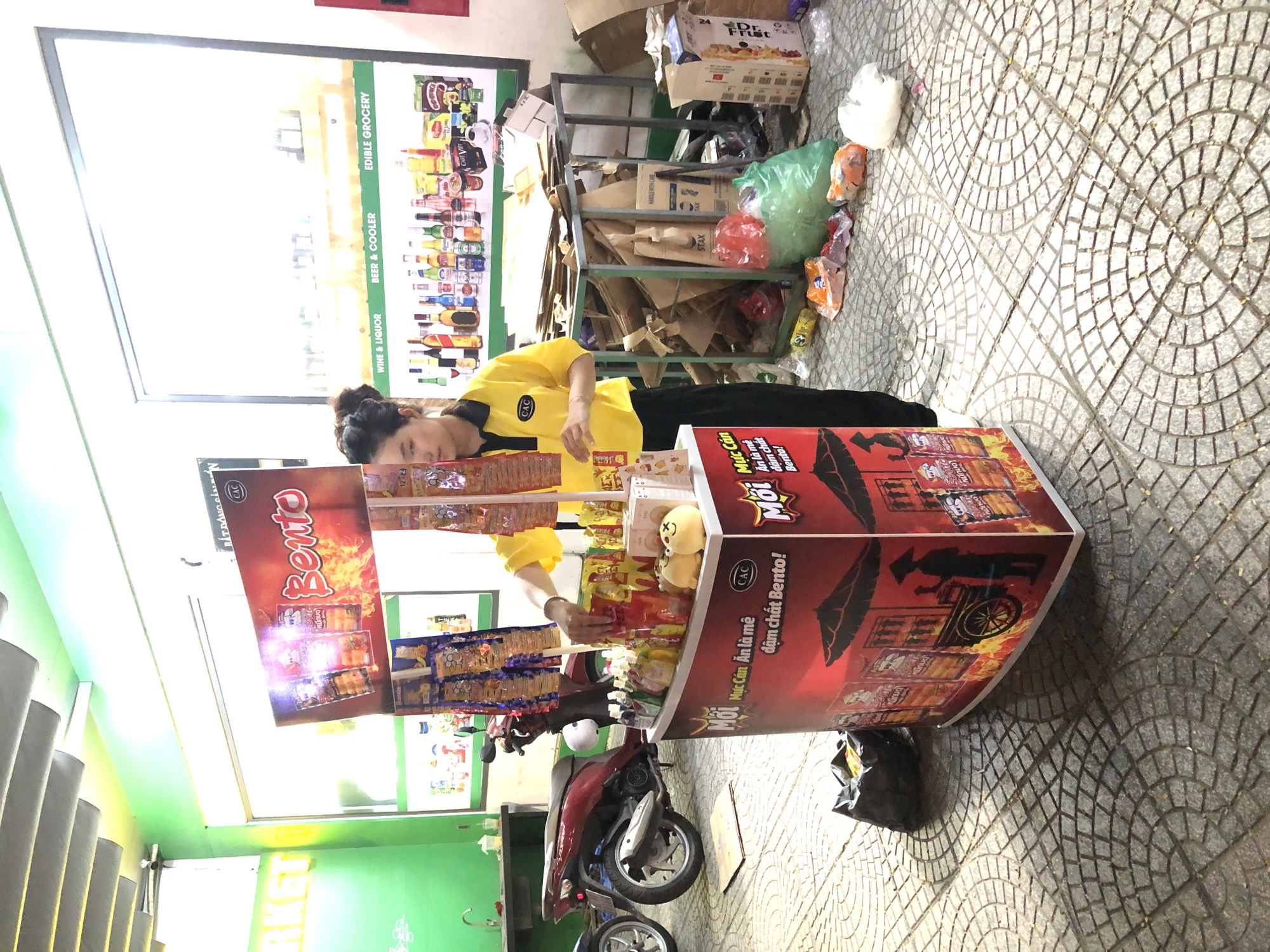 The image shows a street scene in Da Nang, Vietnam. In the foreground, a woman in a yellow polo shirt with a black collar and the CAC logo is standing behind a red and black display stand selling "Bento" snacks. The stand is decorated with images of a street vendor and flames. Several packets of snacks are visible on the stand and hanging from it. To the left of the stand, a red motorcycle is parked. In the background, there is a store with shelves stocked with various products, including bottles of wine and liquor, and packaged foods. A large poster on the store wall advertises "WINE & LIQUOR", "BEER & COOLER", and "EDIBLE GROCERY". There are also stacks of cardboard boxes and bags of trash in the area around the store. The ground is paved with patterned tiles. The overall impression is a bustling street scene in a commercial area.
