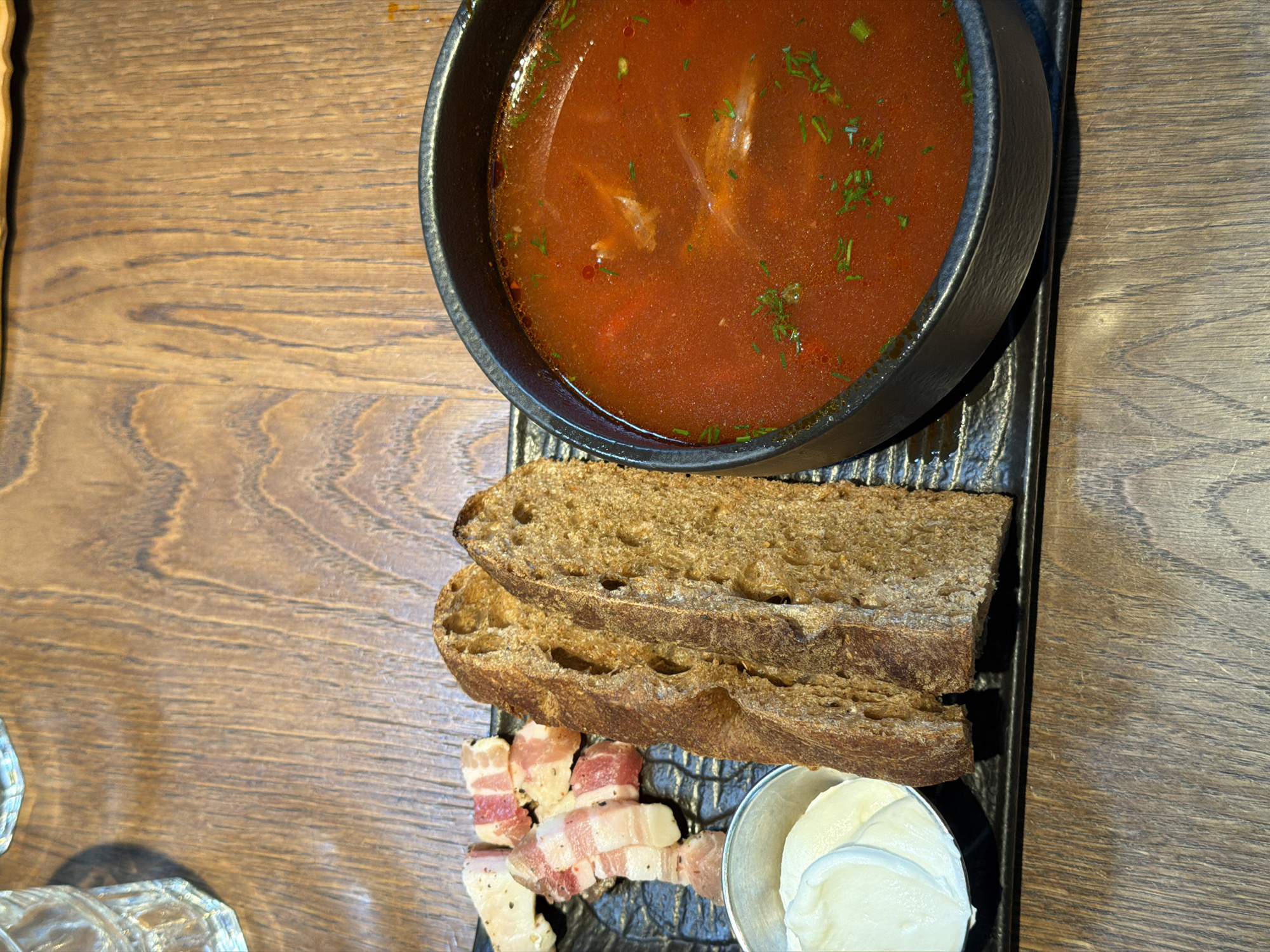 The image displays a traditional Eastern European meal, presented from an overhead perspective on a wooden table. The central item is a dark, round bowl filled with a vibrant reddish-orange soup, likely borscht, generously garnished with chopped green herbs such as dill or parsley.
Alongside the soup, on a textured, dark rectangular serving platter, are several accompaniments. Two thick slices of rustic, dark bread, resembling rye bread, are stacked. Below the bread, several thin slices of cured pork belly or fat, known as salo, are arranged, showing distinct layers of white fat and pinkish meat, seasoned with dark specks of pepper. Next to the salo is a small, silver-toned metal cup holding a portion of a white, creamy condiment, characteristic of sour cream or smetana, often served with borscht.
The setting appears to be indoors, with even lighting illuminating the food and the natural grain of the wooden table. There are no people visible in the image, and no discernible text. The combination of these dishes strongly indicates a traditional meal common in Eastern European cuisine, consistent with the provided location context of Moscow, Russia.