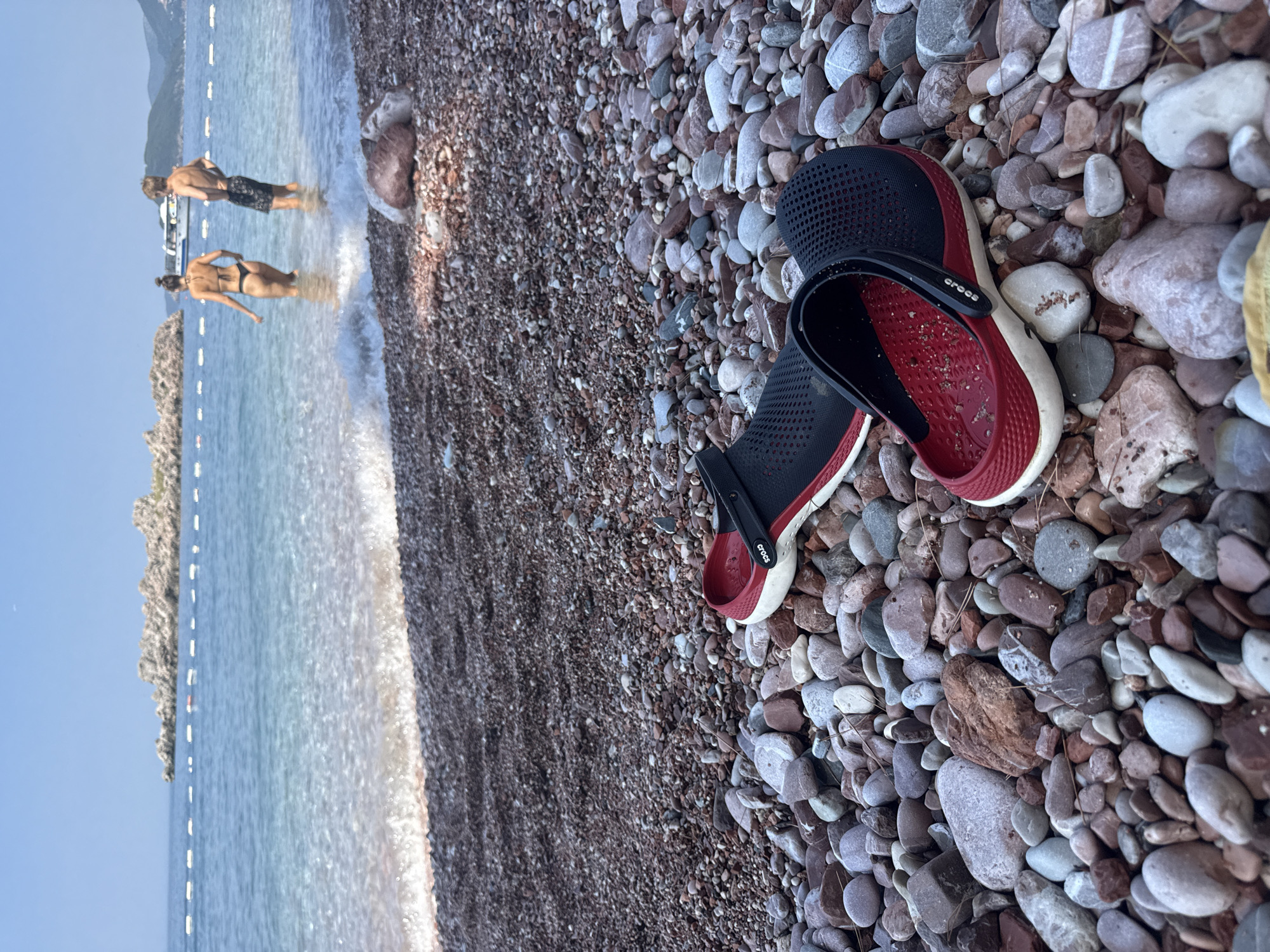 The image shows a close-up of a pair of black and red Crocs resting on a pebble beach. The Crocs are positioned at an angle, with the red interior facing upwards, revealing some sand particles. The beach is composed of a variety of small, smooth pebbles in shades of grey, brown, and red.
In the background, the blue sea gently laps at the shore, creating white foam. The water appears clear and inviting. Further in the distance, two people are visible standing in the shallow water. One person, wearing dark shorts, is facing away from the camera, and the other, wearing a bikini, is also facing away, with their back to the viewer. Their figures are somewhat blurred due to the distance and the focus on the foreground objects.
The scene suggests a sunny day at a beach location, likely in Bečići, Montenegro, given the context. The presence of people in swimwear and the tranquil sea indicate a recreational setting. The overall mood conveyed is one of relaxation and enjoyment of a summer day by the water. The Crocs suggest a casual approach to beachwear. The sky is a clear, bright blue. The shoreline is marked by a string of buoys or markers.